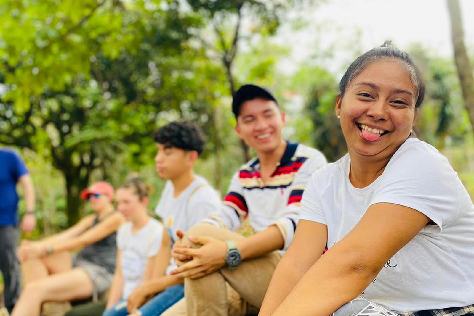 A group of young people is gathered outdoors, some sitting on a bench and others sitting on the ground. The person in the foreground, wearing a white t-shirt, is smiling and sticking out their tongue with trees visible in the background.