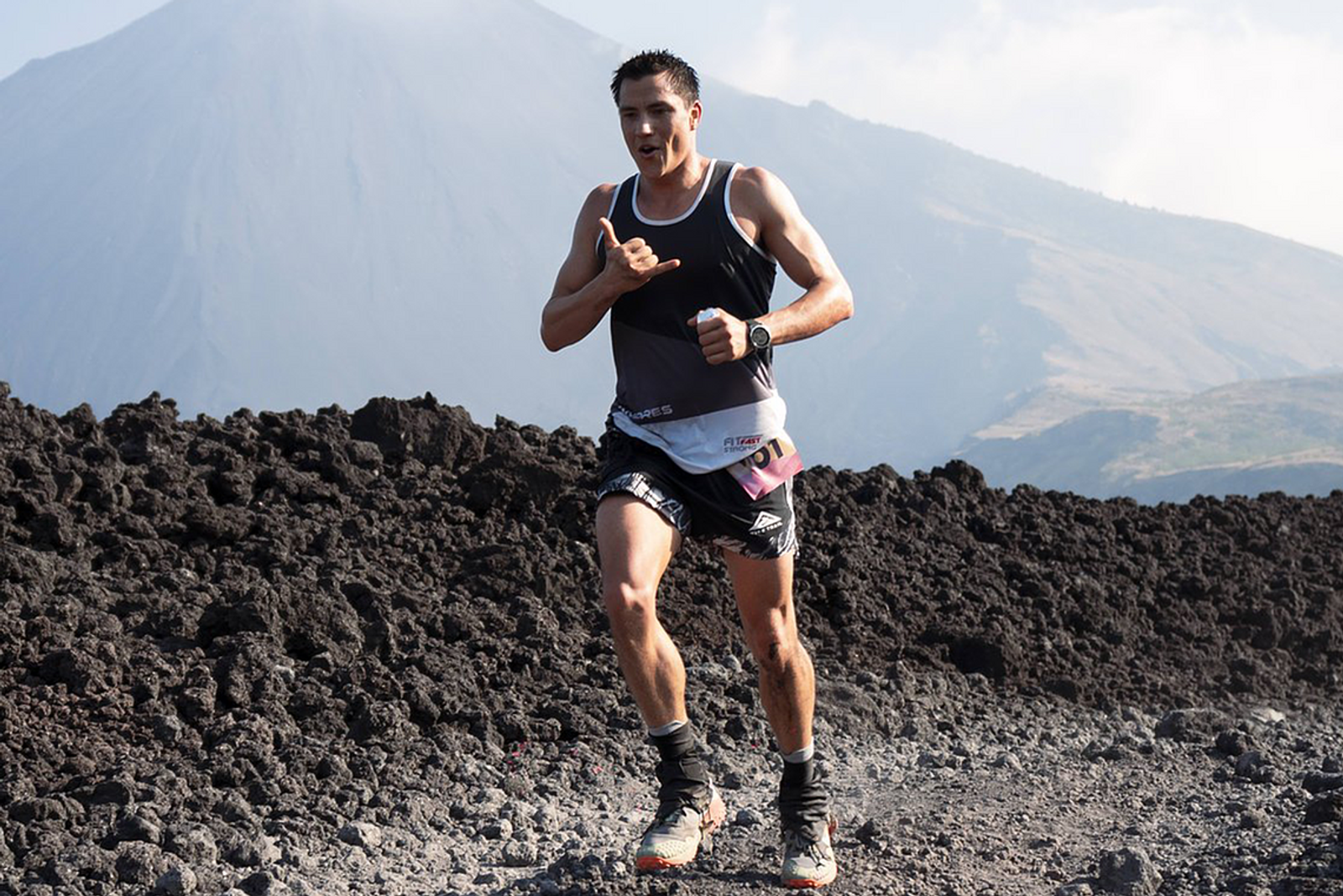A person wearing athletic gear is running on a rocky path with a mountainous background. The runner appears determined, and the landscape includes rough volcanic terrain. The sky is clear with a few clouds.