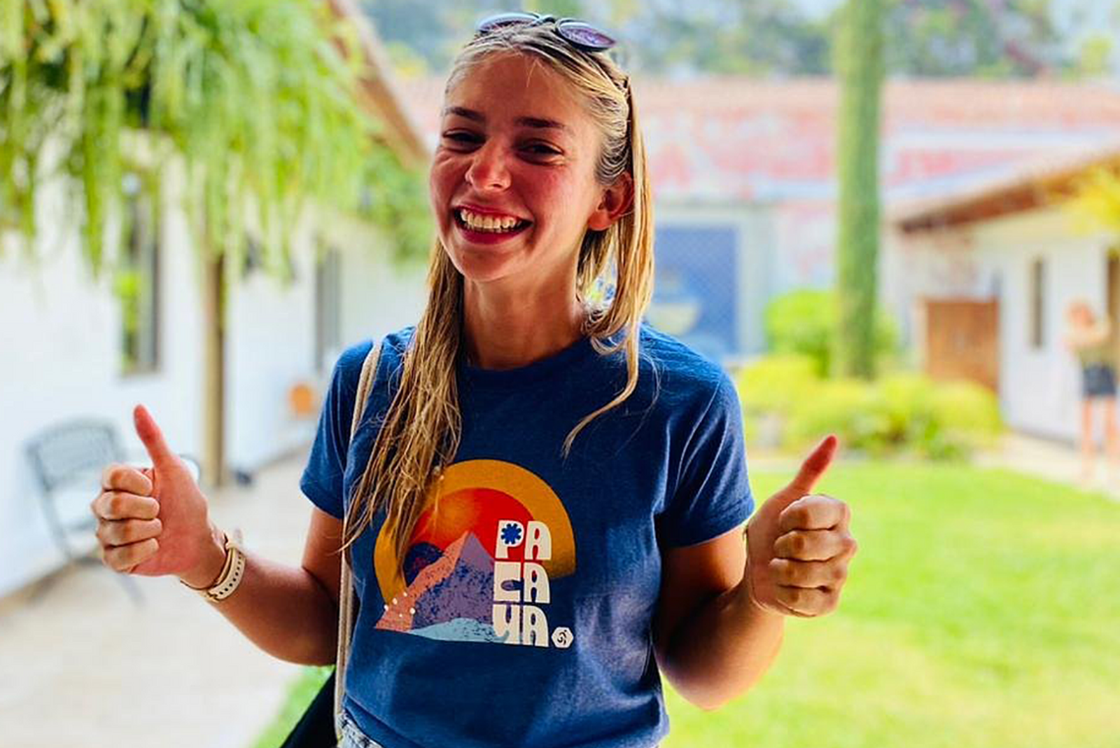 A woman stands outdoors, smiling broadly and giving two thumbs up. She is wearing a blue t-shirt with a colorful graphic design on it. A building and greenery are visible in the background. She has long blonde hair and is wearing sunglasses on her head.