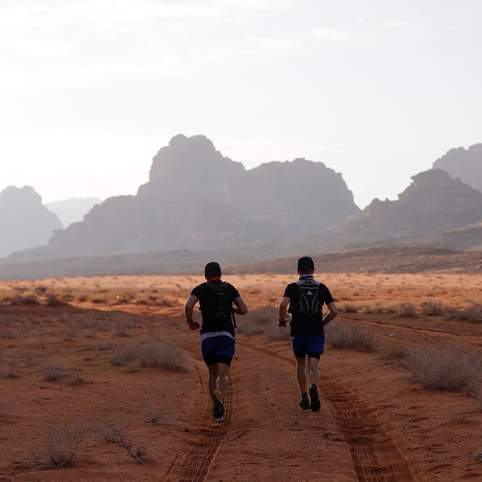 Two people run along a desert trail with red, sandy terrain. They wear black shirts, shorts, backpacks, and caps. In the background, eroded rock formations rise under a hazy sky. It is early morning or late afternoon, evident from the soft sunlight.