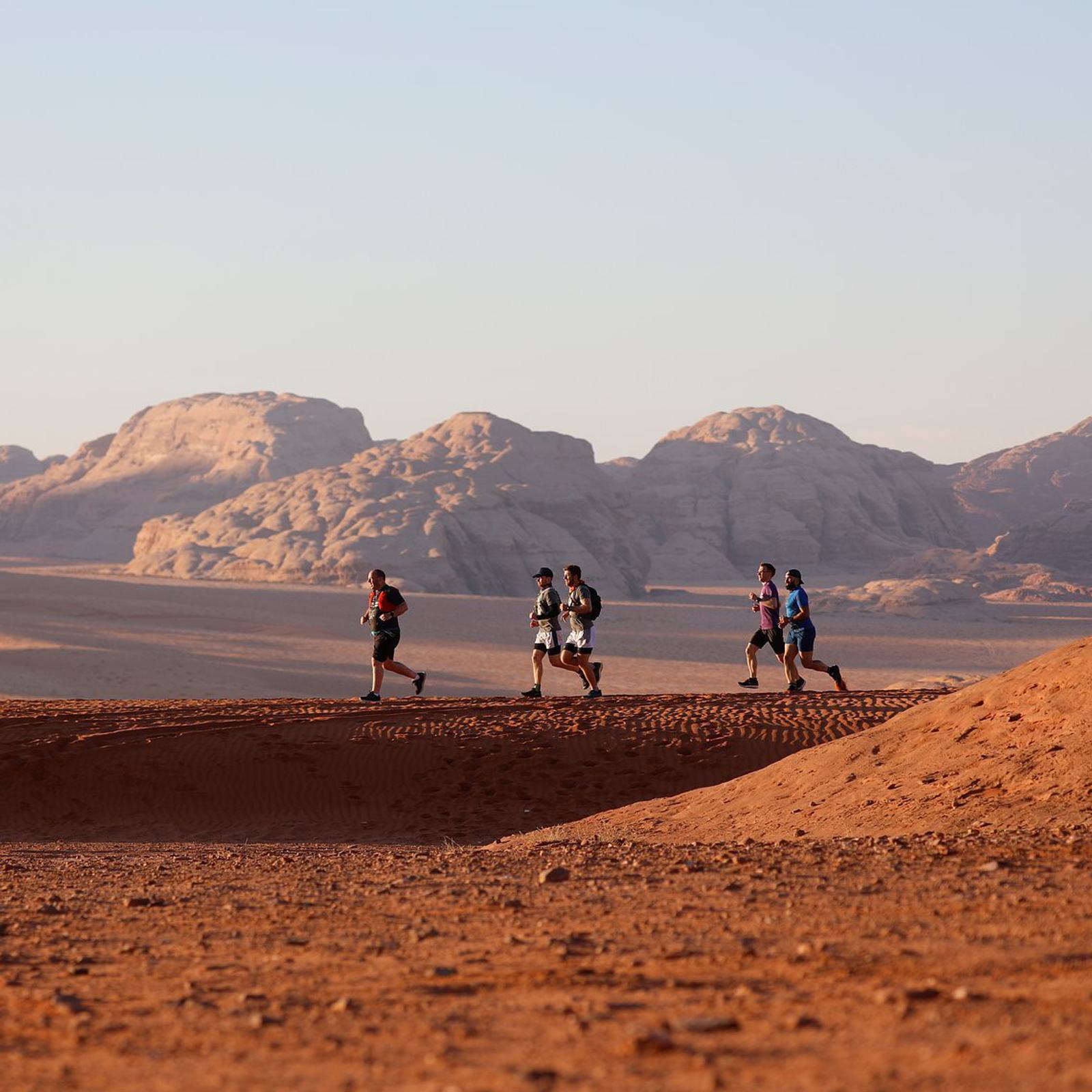 Four runners in athletic gear are running on a sandy trail in a desert landscape. The background features vast, rocky formations and a clear sky, suggesting a remote and challenging environment. The scene is bathed in soft, natural light.