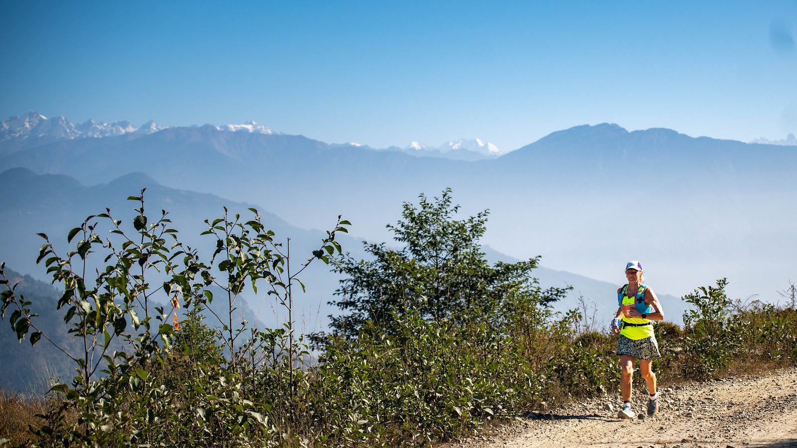 A runner wearing a green and yellow top, shorts, and a white cap is jogging along a dirt trail surrounded by bushes. Behind them, the landscape features rolling hills and towering mountain peaks under a clear, blue sky.
