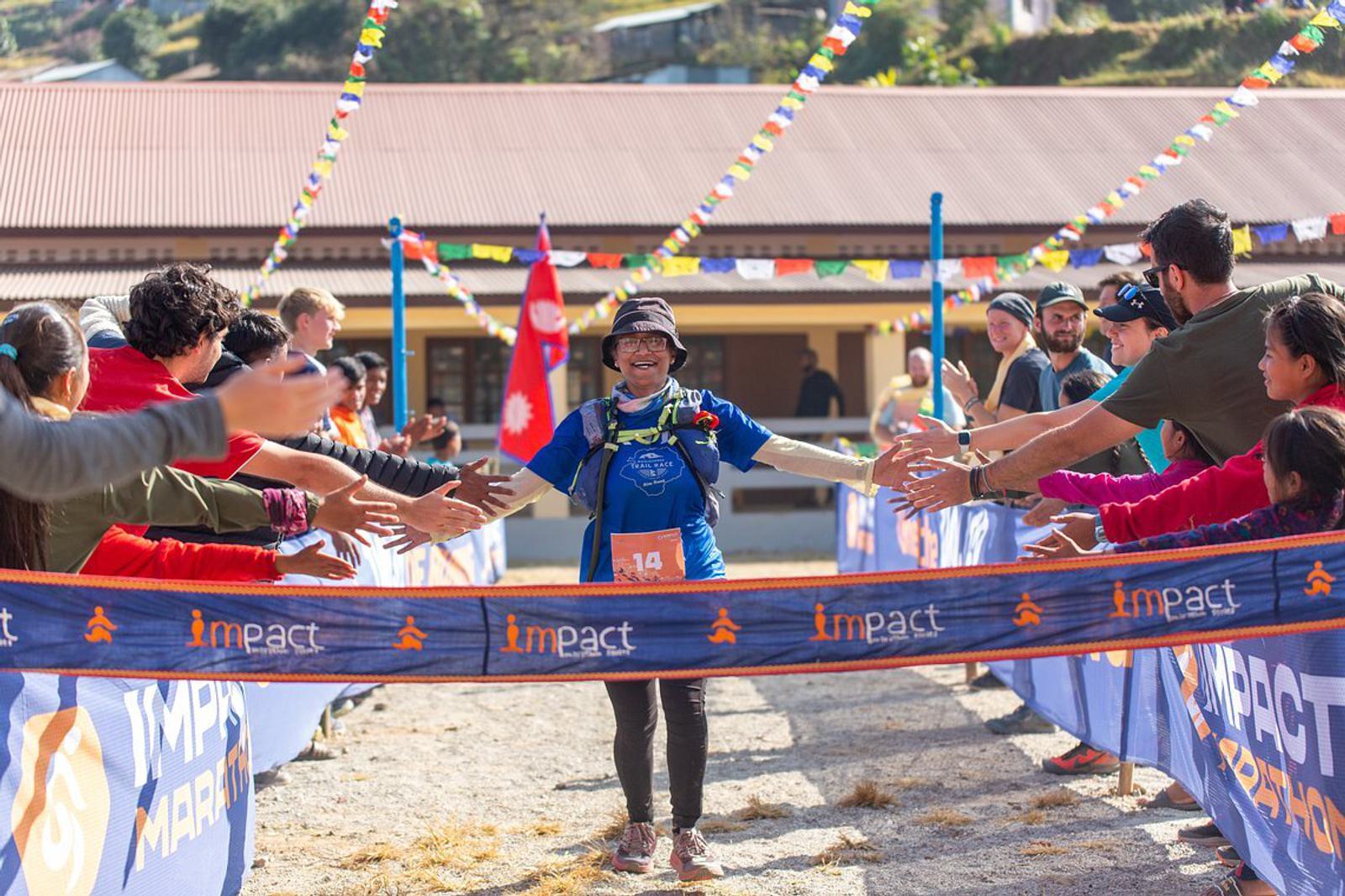 A runner wearing a blue shirt and cap is crossing the finish line at a race event. She is smiling and high-fiving spectators standing on both sides of the path. Colorful flags and a building with a corrugated roof are visible in the background.