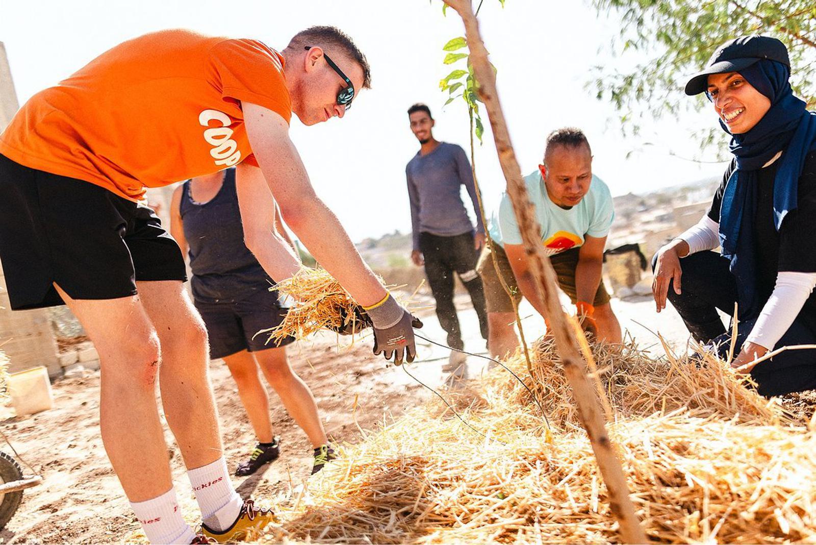 A group of people is working together outdoors, smiling as they handle straw or hay. The participants, diverse in gender and attire, are engaged in a community or environmental project under sunny, clear conditions. Nearby trees and bright daylight are visible.