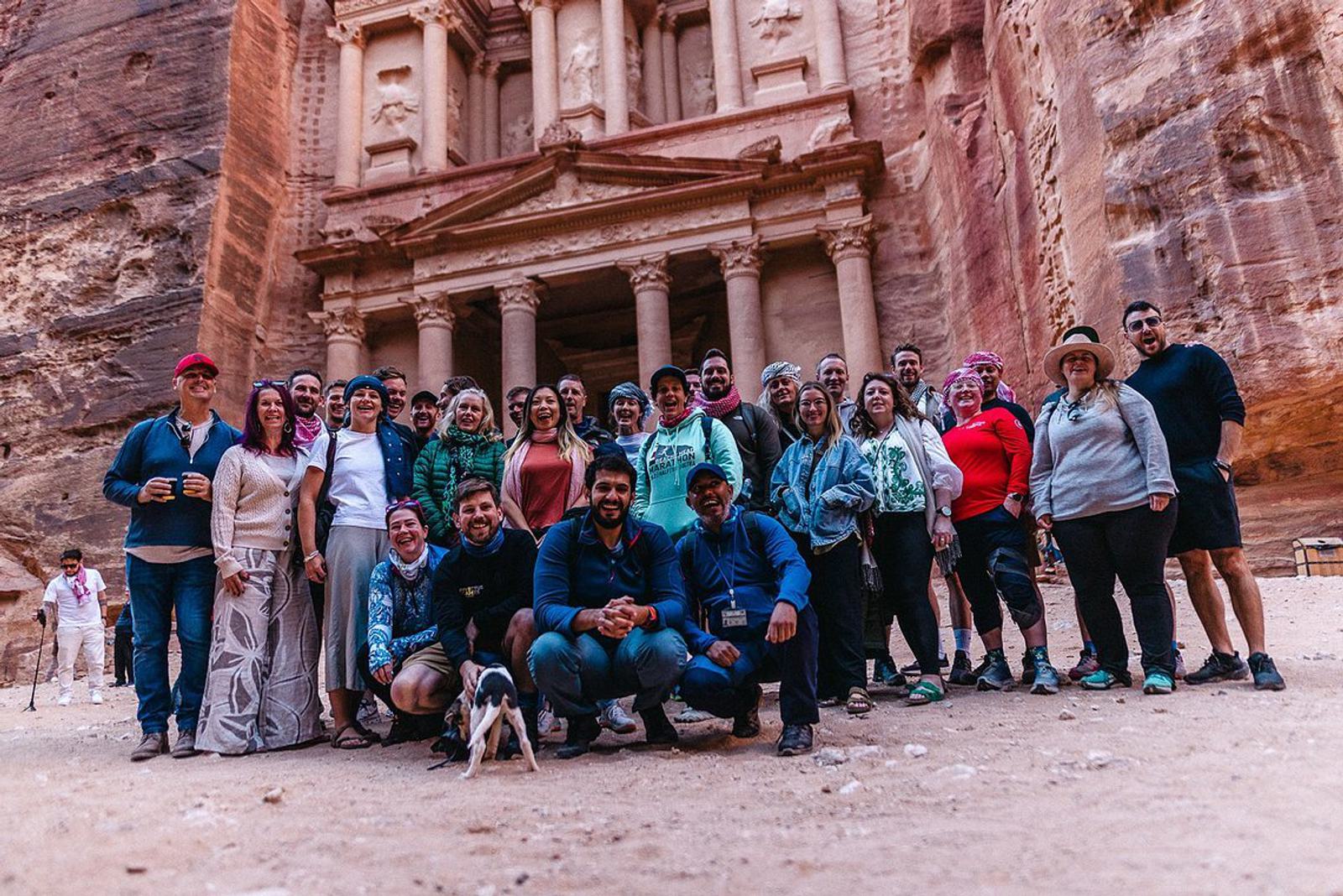 A diverse group of people pose in front of the ancient rock-cut architecture of Petra in Jordan. The group is smiling and dressed casually, with some members wearing hats and sunglasses. The monumental sandstone facade towers behind them.
