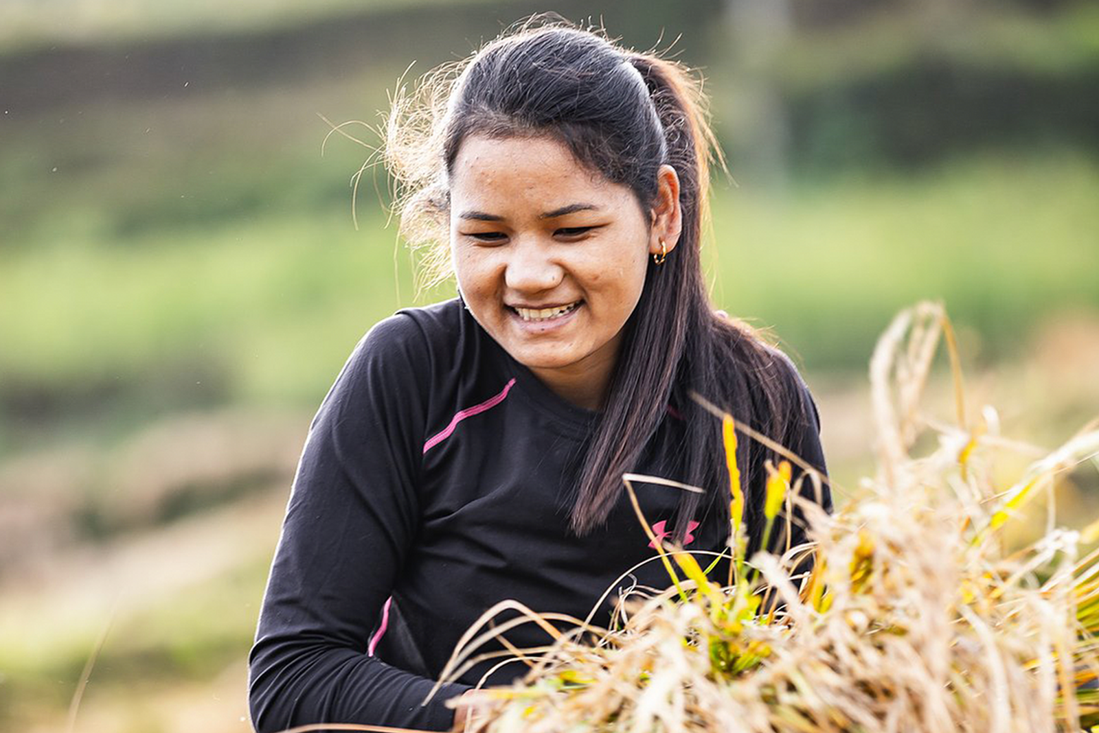 A young woman with long dark hair and wearing a black shirt sits outdoors in a field, smiling while holding what appears to be a bundle of harvested crops. The background is green and blurred, indicating a rural or agricultural setting.