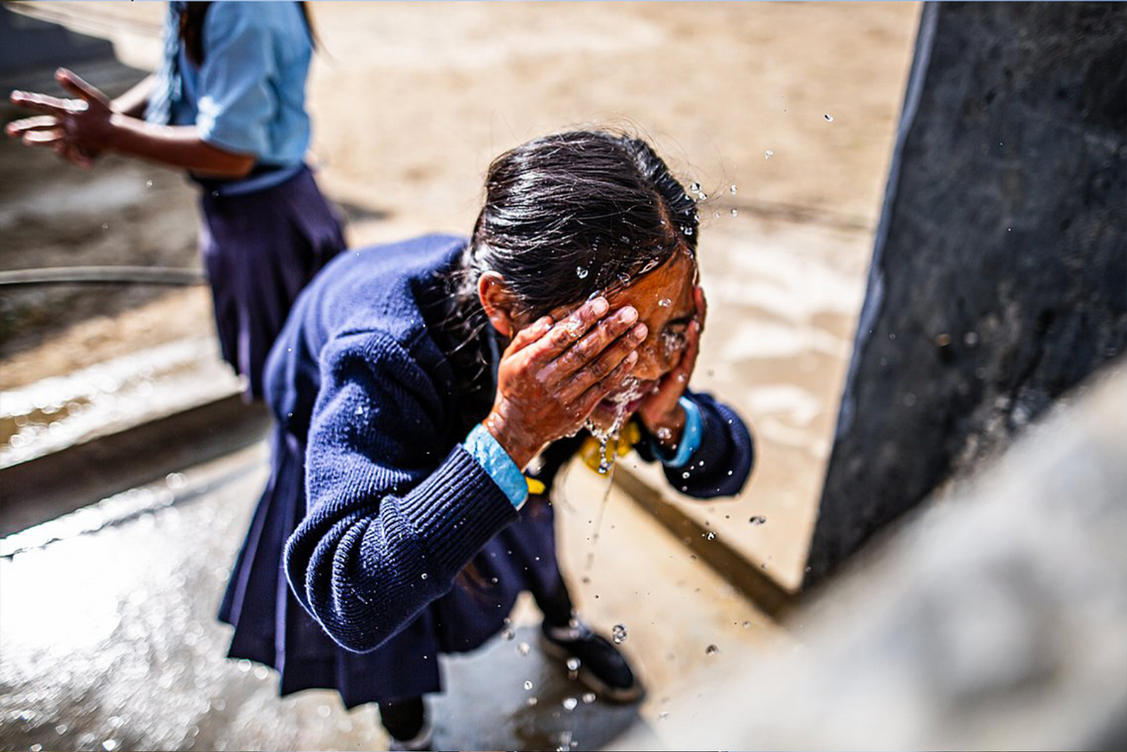 A young girl wearing a navy blue sweater and a skirt splashes water on her face from a spout outdoors. Another person is partially visible in the background. The surroundings appear to be a mix of concrete and dirt ground.
