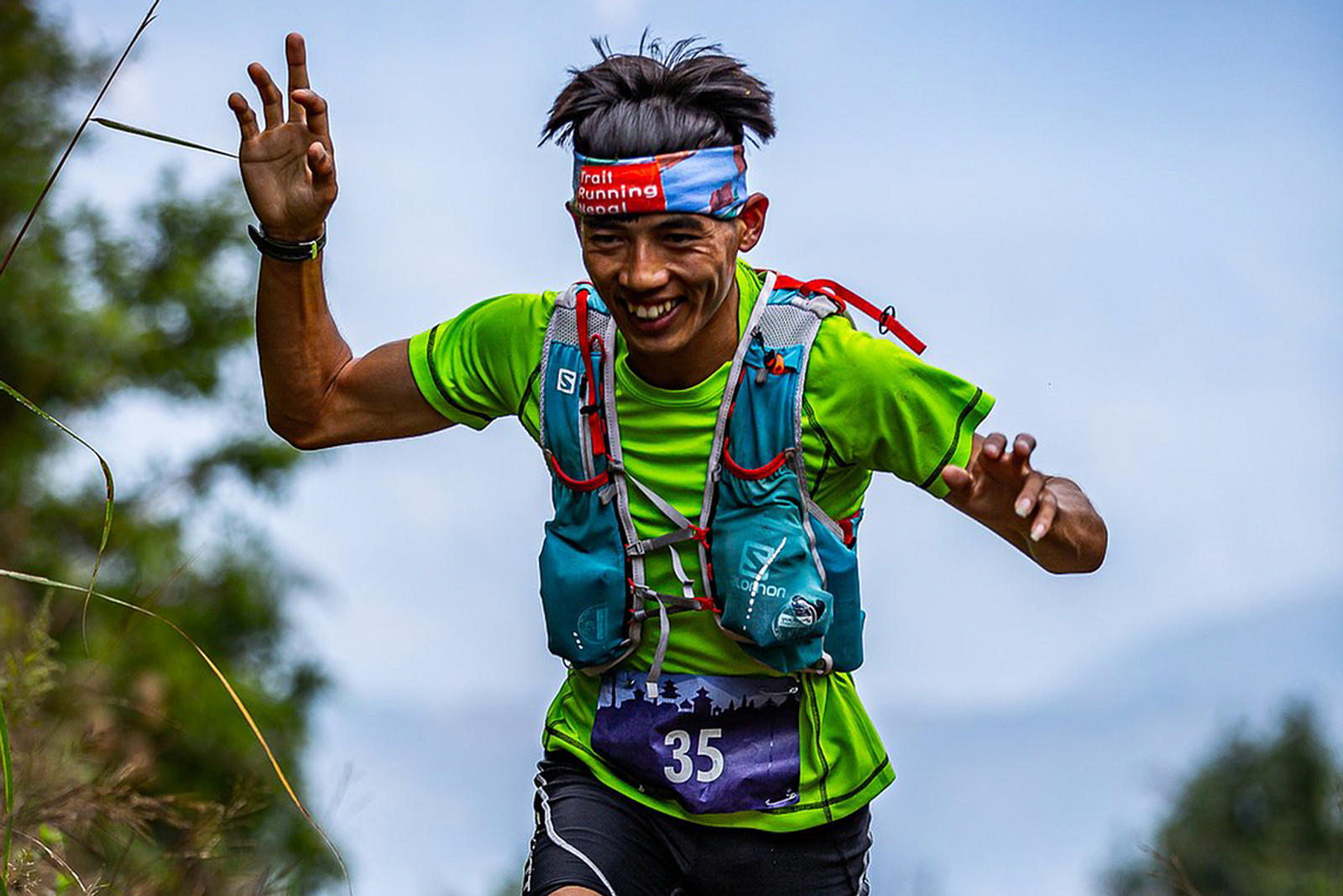 A person wearing a green shirt, blue hydration vest, and headband runs on an outdoor trail, smiling energetically with both hands raised. The background features blurred greenery and a clear sky, suggesting a nature setting. The runner has race number 35 on their shorts.