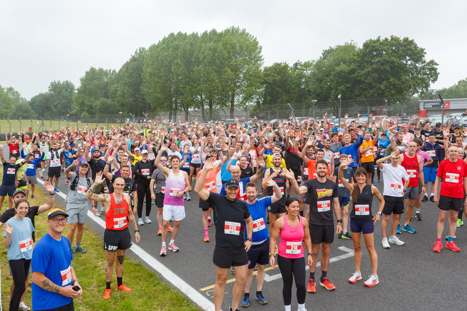 A large group of people in running gear gather on a race track, smiling and waving. The participants hold numbered bibs. Green trees are visible in the background under a cloudy sky.