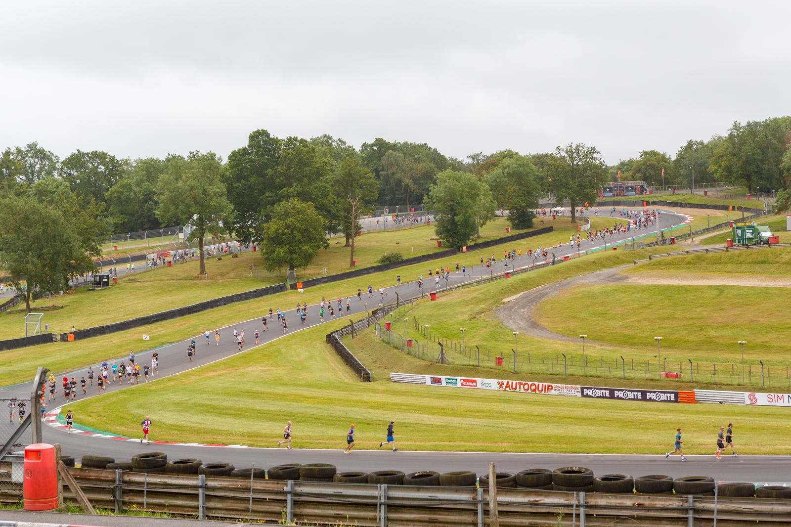 A group of runners participating in a race on a curving track surrounded by green grass and trees, under a cloudy sky. Barricades and advertisements line the track. Spectators can be seen in the background.