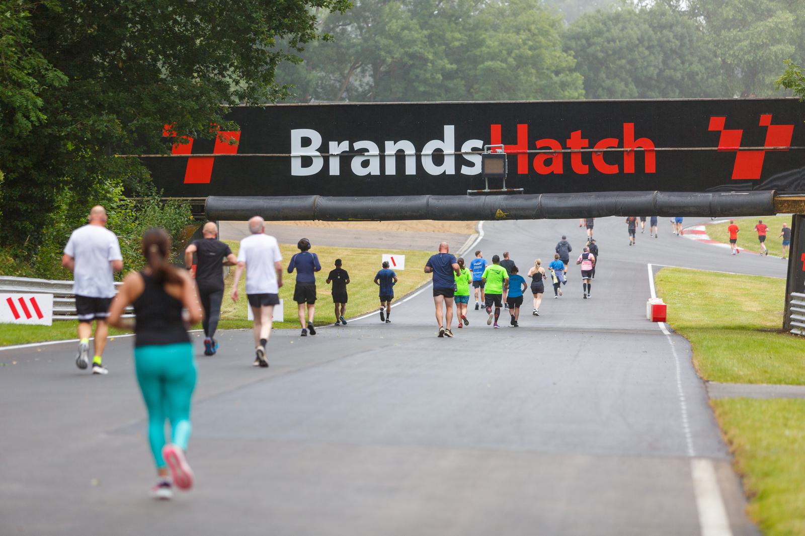People are jogging and walking on a paved track under a "Brands Hatch" sign. The scene is outdoors with greenery and trees lining the path.