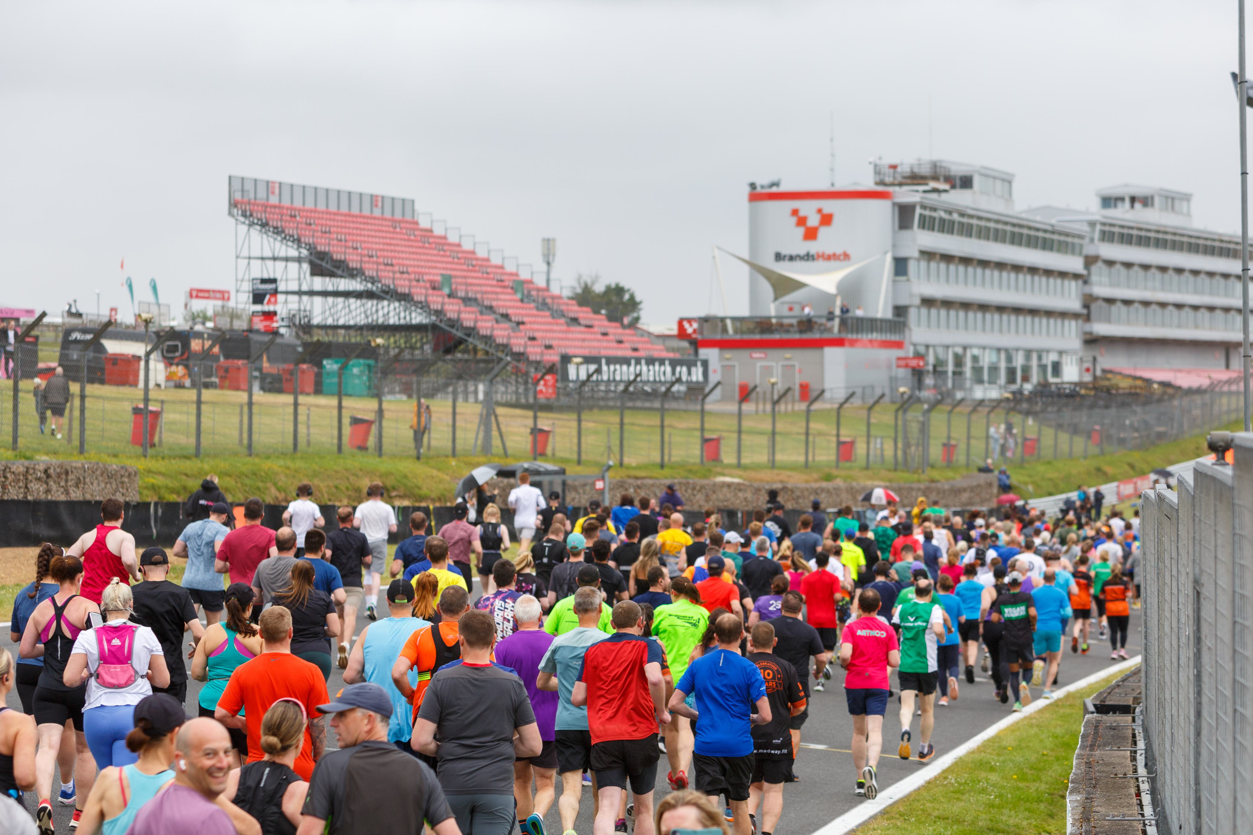 A large group of runners participate in a race on a track. The crowd is diverse, with participants wearing colorful athletic clothing. In the background, there's a grandstand and a building with "Brands Hatch" signage. The sky is overcast.