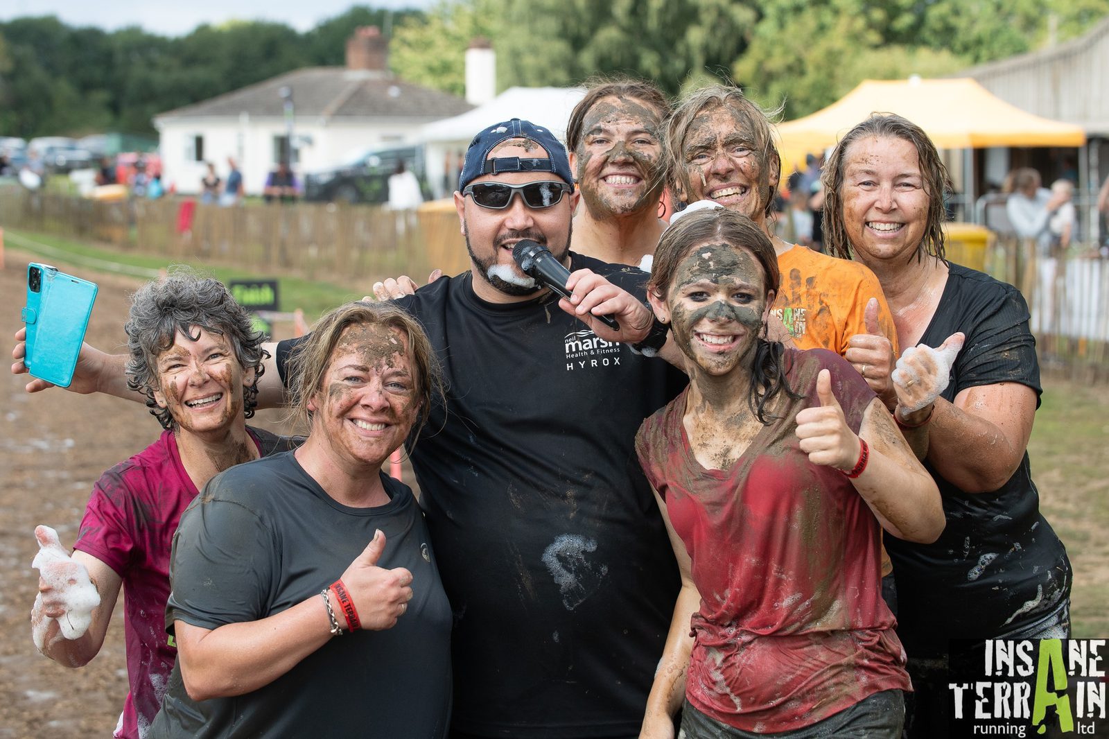A group of six muddy participants pose for a photo after an obstacle course race. One man in the middle holds a microphone and wears sunglasses. The women around him smile and give thumbs up. The event branding "Insane Terrain" is visible in the bottom right corner.