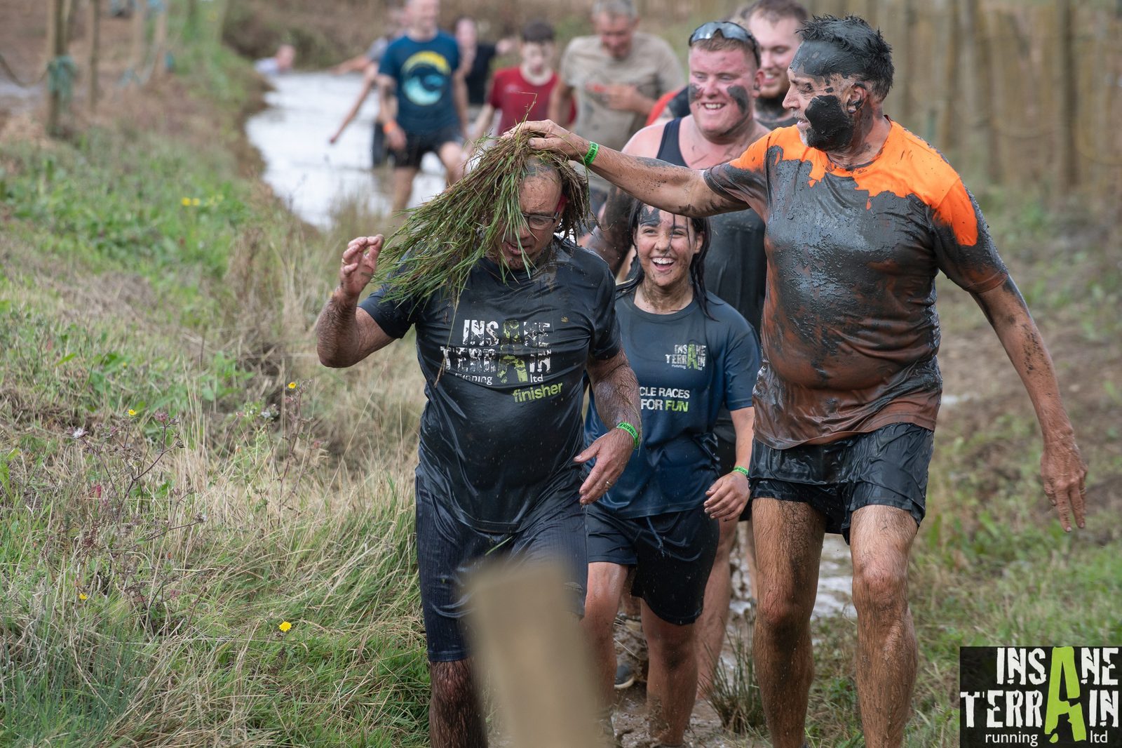 A group of people participates in an outdoor mud run event. One person playfully places a bunch of grass on another's mud-covered head. They all wear athletic clothing, and their expressions show a mix of exhaustion and joy. The "Insane Terrain" logo is visible in the foreground.
