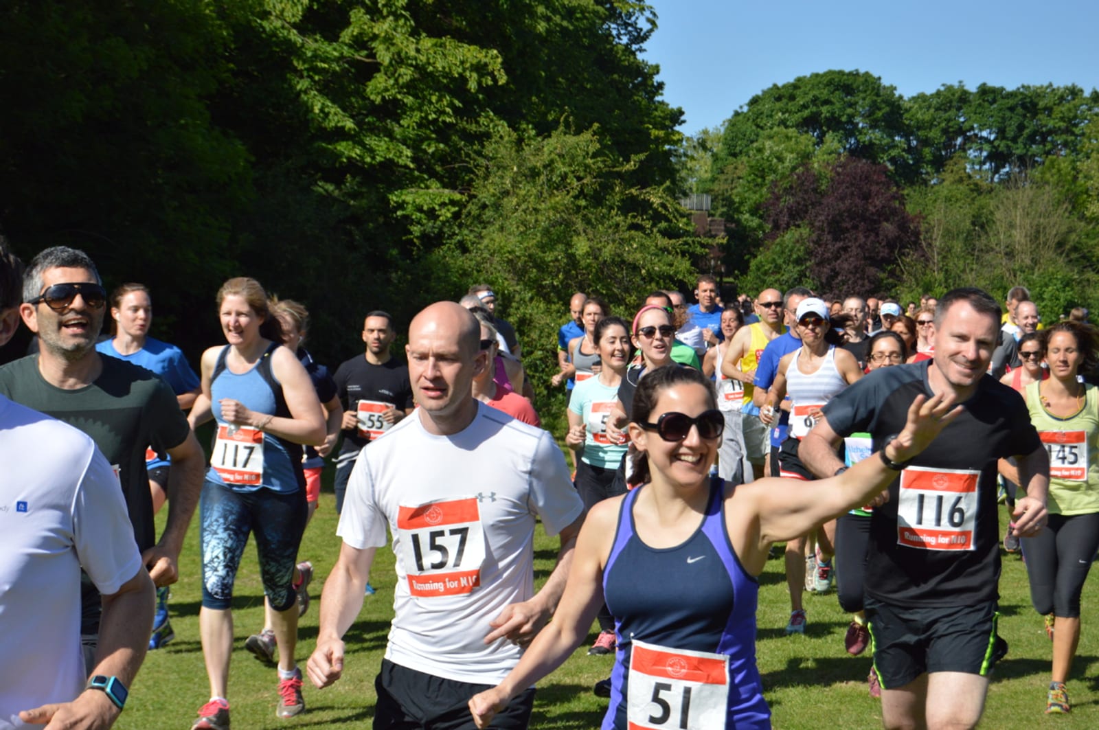 A group of people running a race in a park on a sunny day. They are wearing race bibs with numbers, athletic clothing, and some are smiling and cheering. The background features trees and greenery.