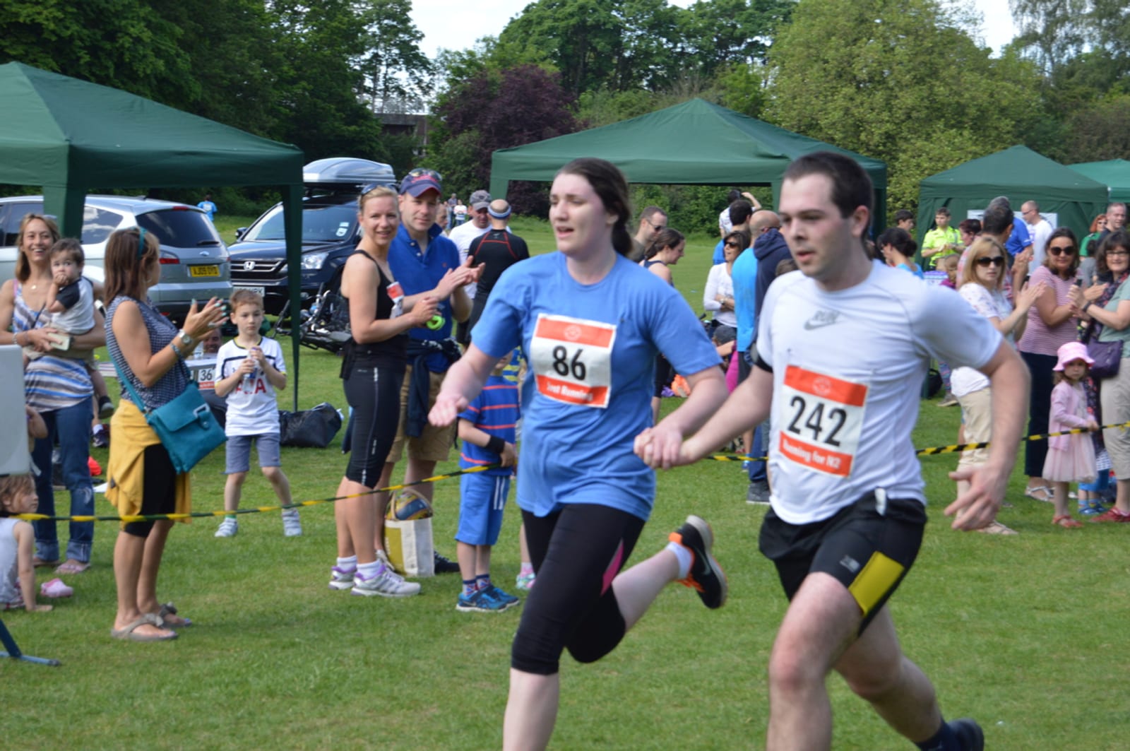 Runners in a race on a grassy field, surrounded by spectators and green tents. Two participants, wearing bib numbers 86 and 242, are running side by side while an audience claps and cheers them on. Outdoor event on a sunny day with trees in the background.
