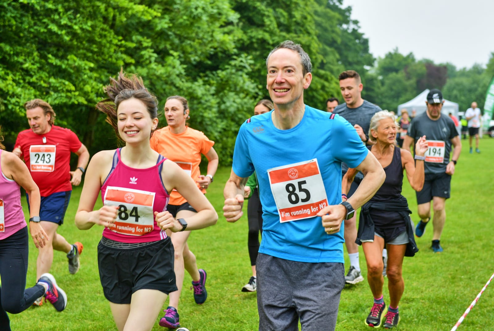 A group of runners, wearing numbered bibs, participates in an outdoor race. The man (85) and woman (84) in the foreground are smiling and jogging side by side. Other runners are visible in the background, and they are surrounded by greenery on an overcast day.