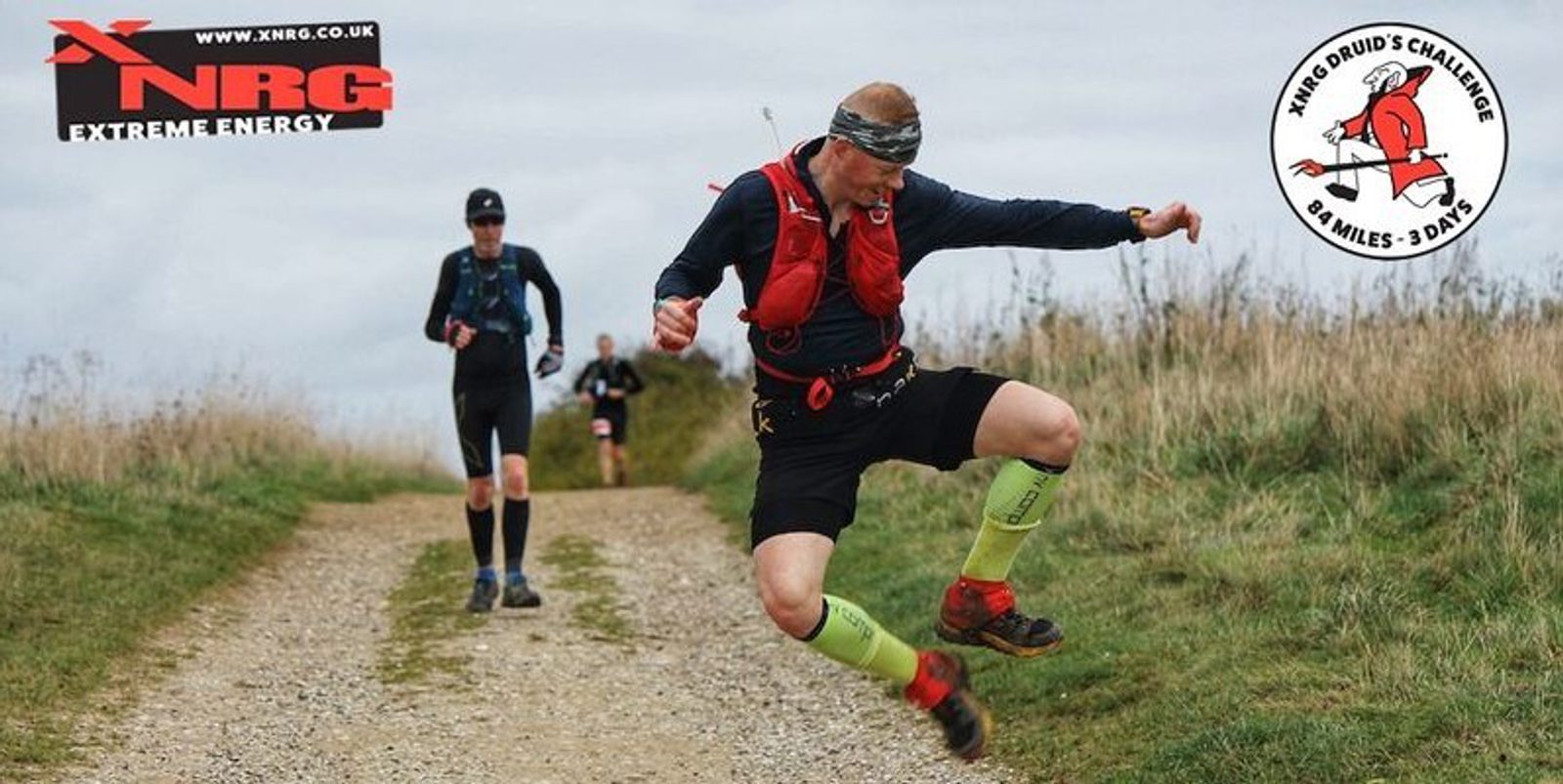 A trail runner in a red vest and green socks jumps mid-air on a gravel path. Another runner follows behind. The logos of XNRG Extreme Energy and XNRG Druid's Challenge 84 Miles - 3 Days are displayed in the top left and right corners, respectively.