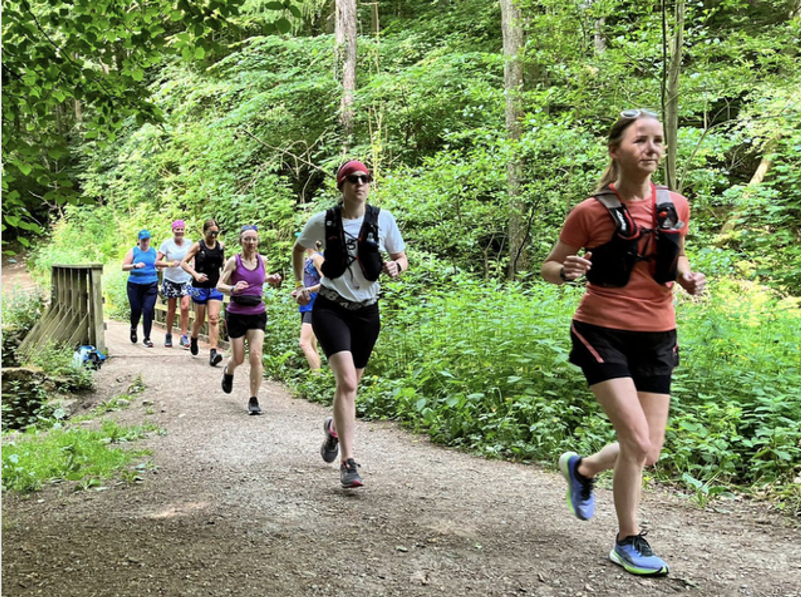 A group of runners jogs along a forest trail during the day. The path is surrounded by lush greenery and tall trees. The runners are dressed in athletic gear, with most wearing shorts and t-shirts or tank tops. Several are equipped with hydration packs.