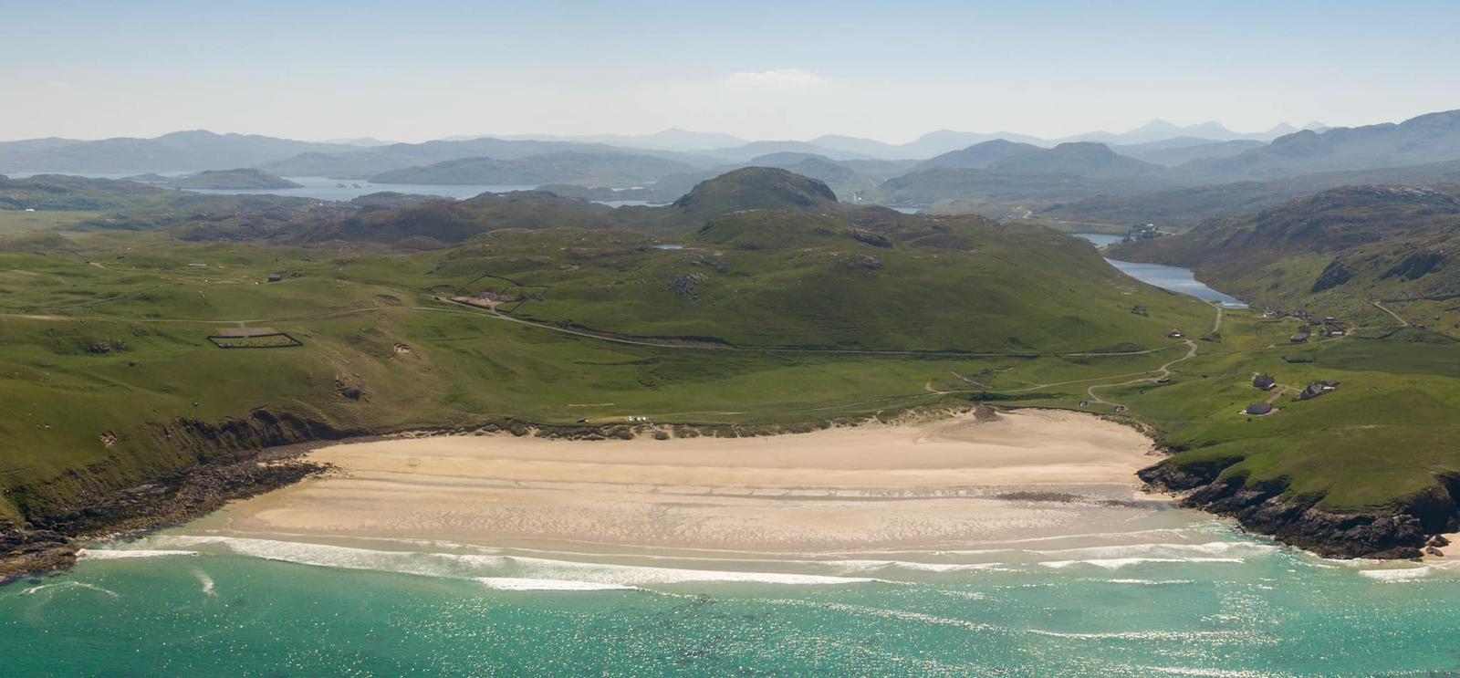 Aerial view of a serene beach with turquoise waters, rolling waves, and golden sand, backed by vast green hills. Dotted throughout the landscape are clusters of small houses and winding roads, set against a backdrop of hazy mountains under a clear sky.