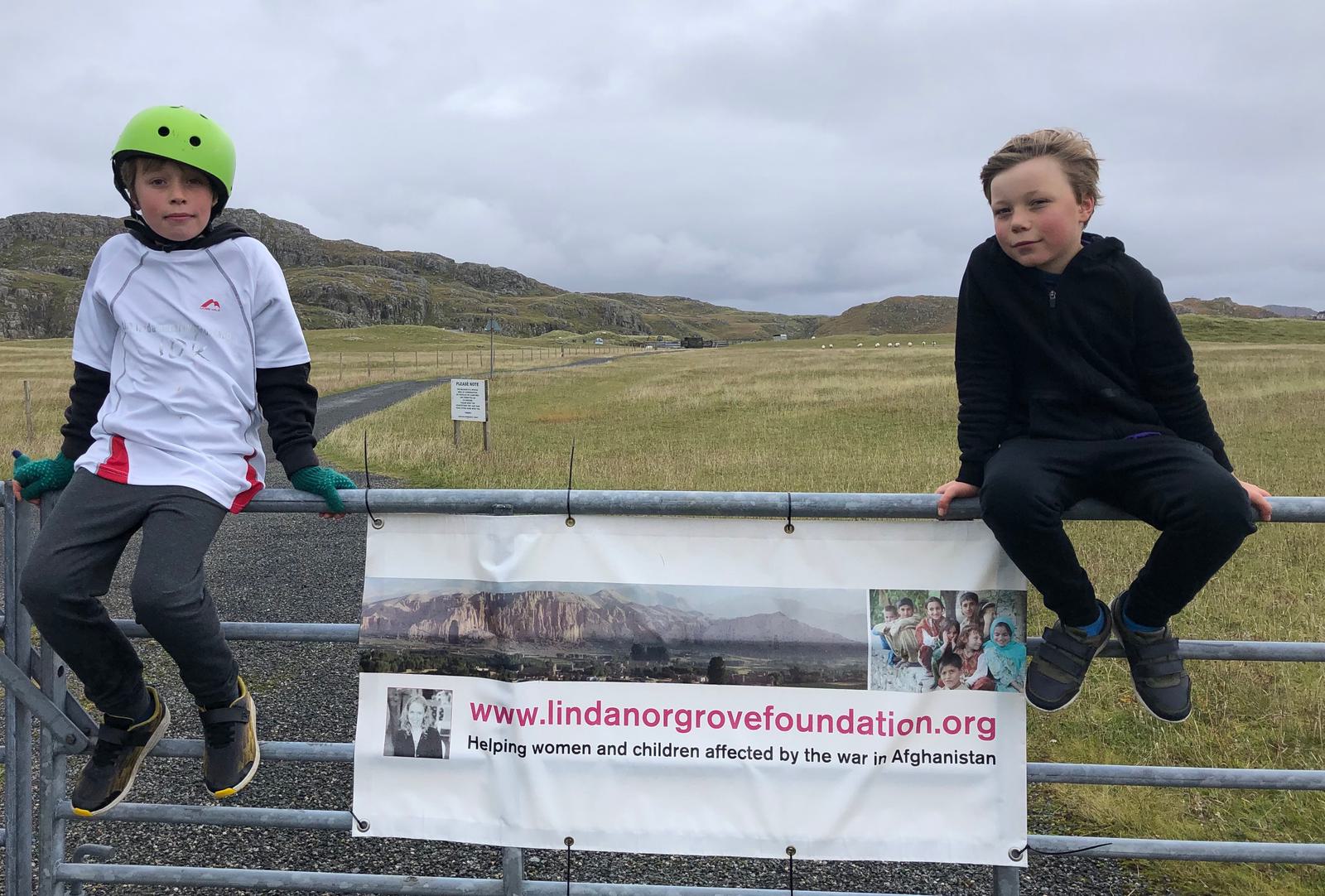 Two children sit on a metal gate in a grassy area with hills in the background. One child wears a helmet and gloves, and the other wears a black jacket. They are near a banner for the Linda Norgrove Foundation, which helps women and children affected by the war in Afghanistan.