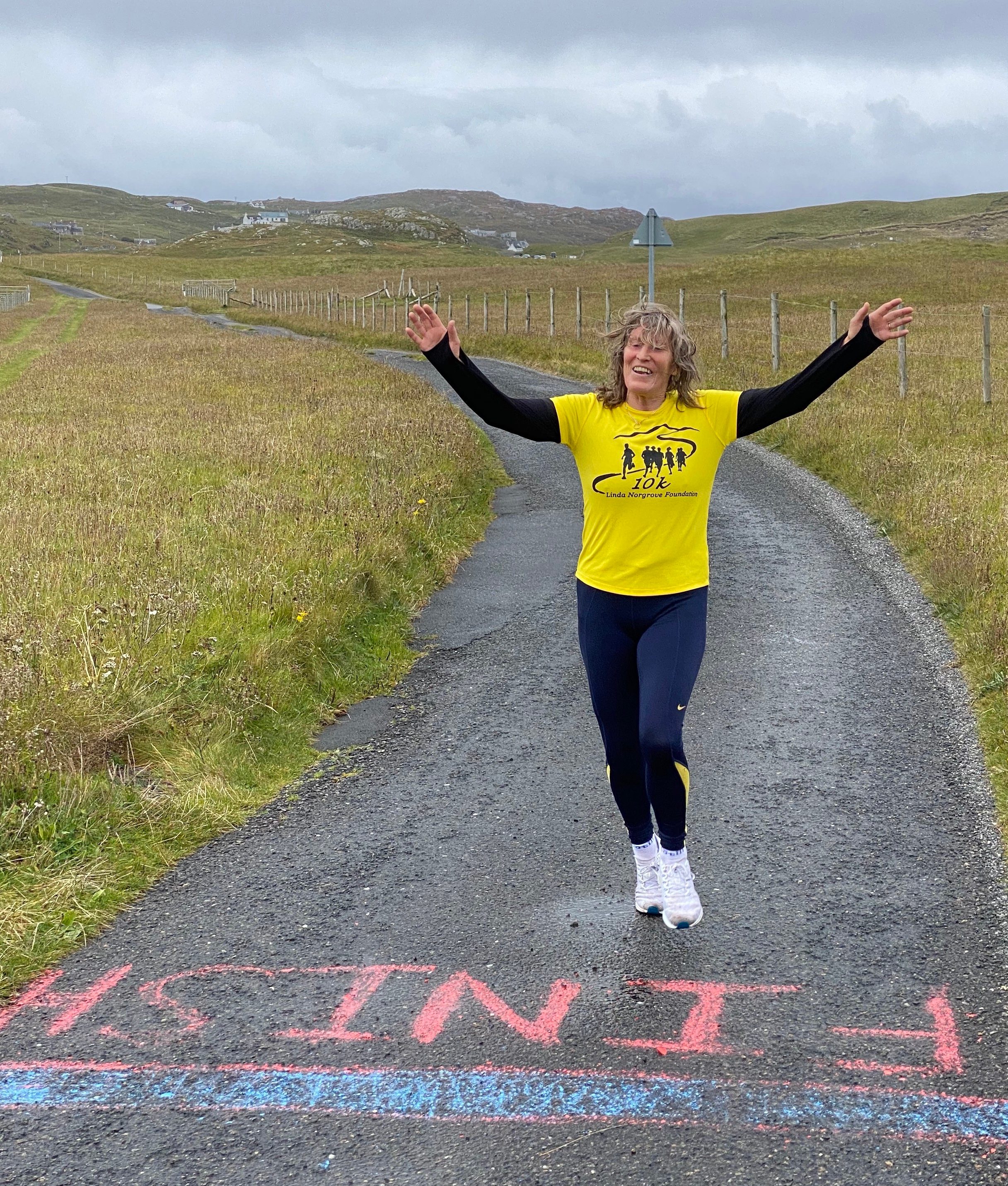 A person wearing a yellow shirt and blue leggings raises their arms in celebration as they cross a finish line painted on a rural road. Grassy fields, hills, and cloudy skies are in the background.