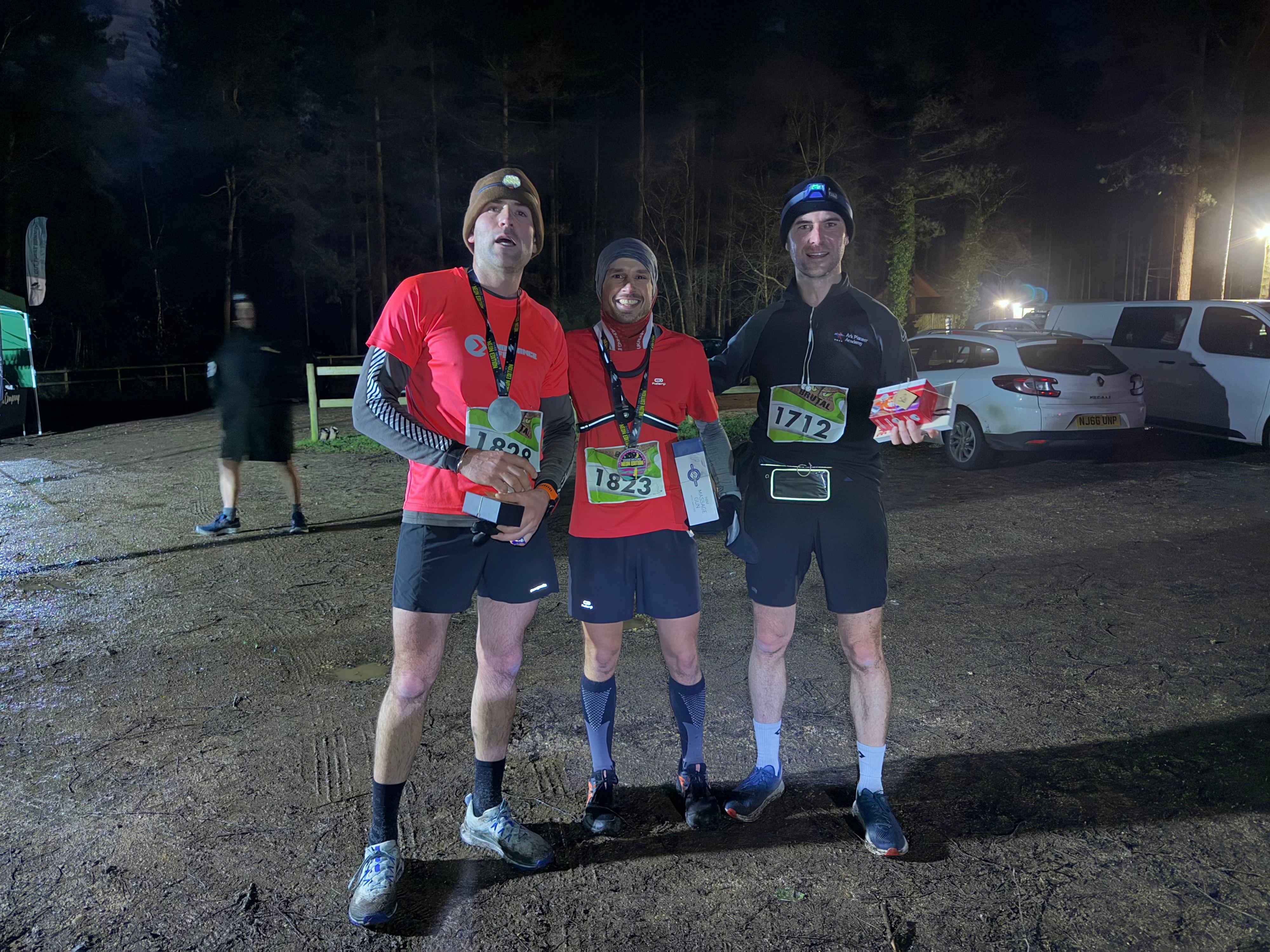 Three runners posing outdoors at night, wearing race bibs and athletic clothing. The area is dimly lit with parked cars in the background. One person is holding a small box.