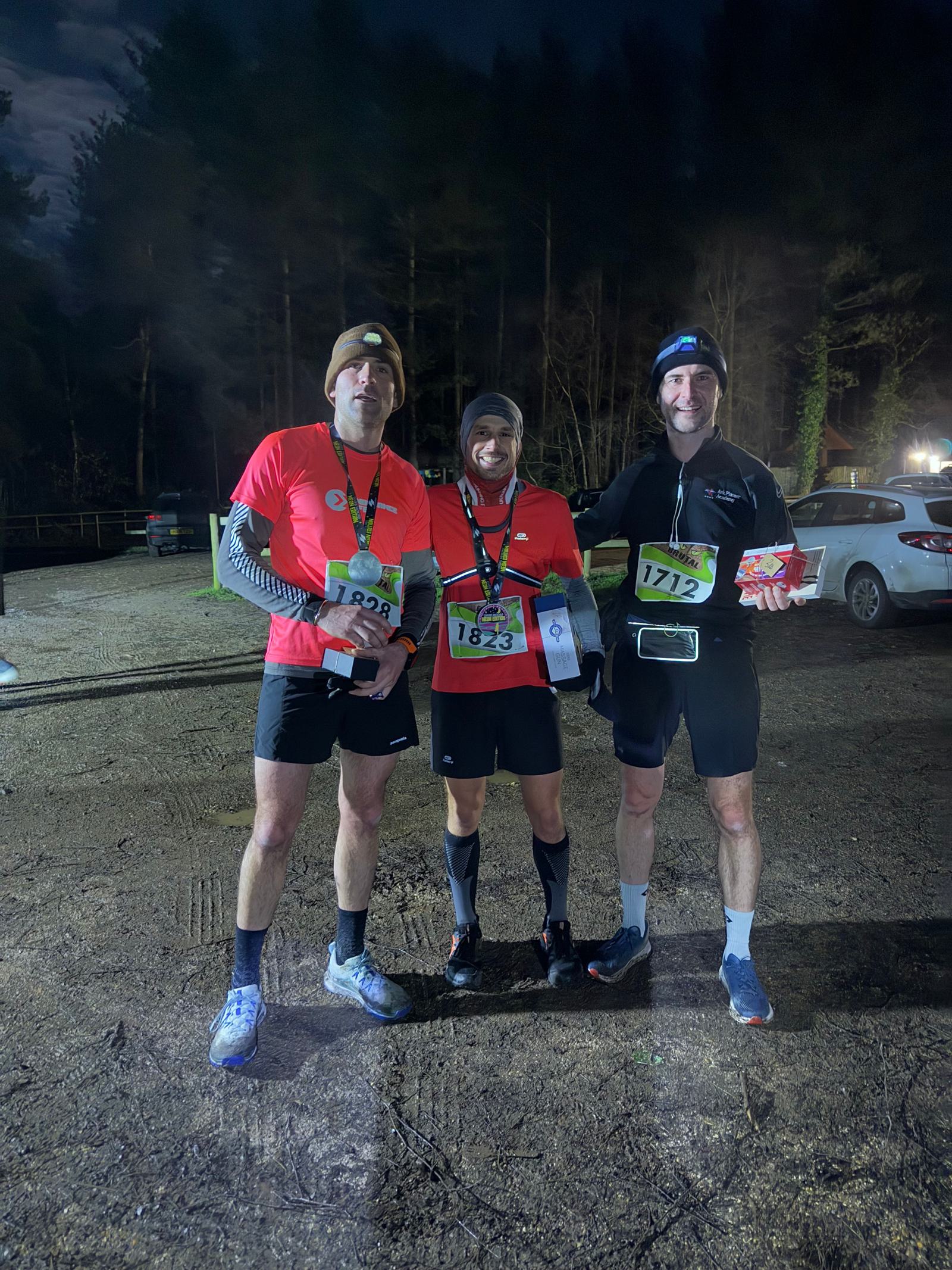 Three runners wearing race bibs stand together in a dimly lit outdoor area. Two hold prizes and all wear winter running gear. Trees and parked cars are visible in the background.
