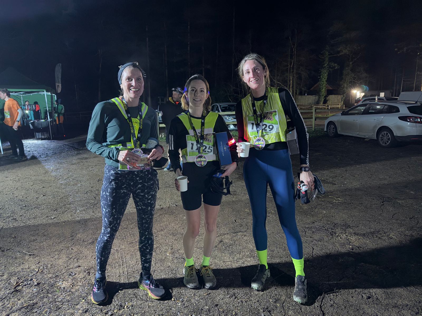 Three people in athletic gear stand outdoors at night, smiling and wearing medals. They each have race bibs and are holding refreshments. A lit tent and parked cars are visible in the background.