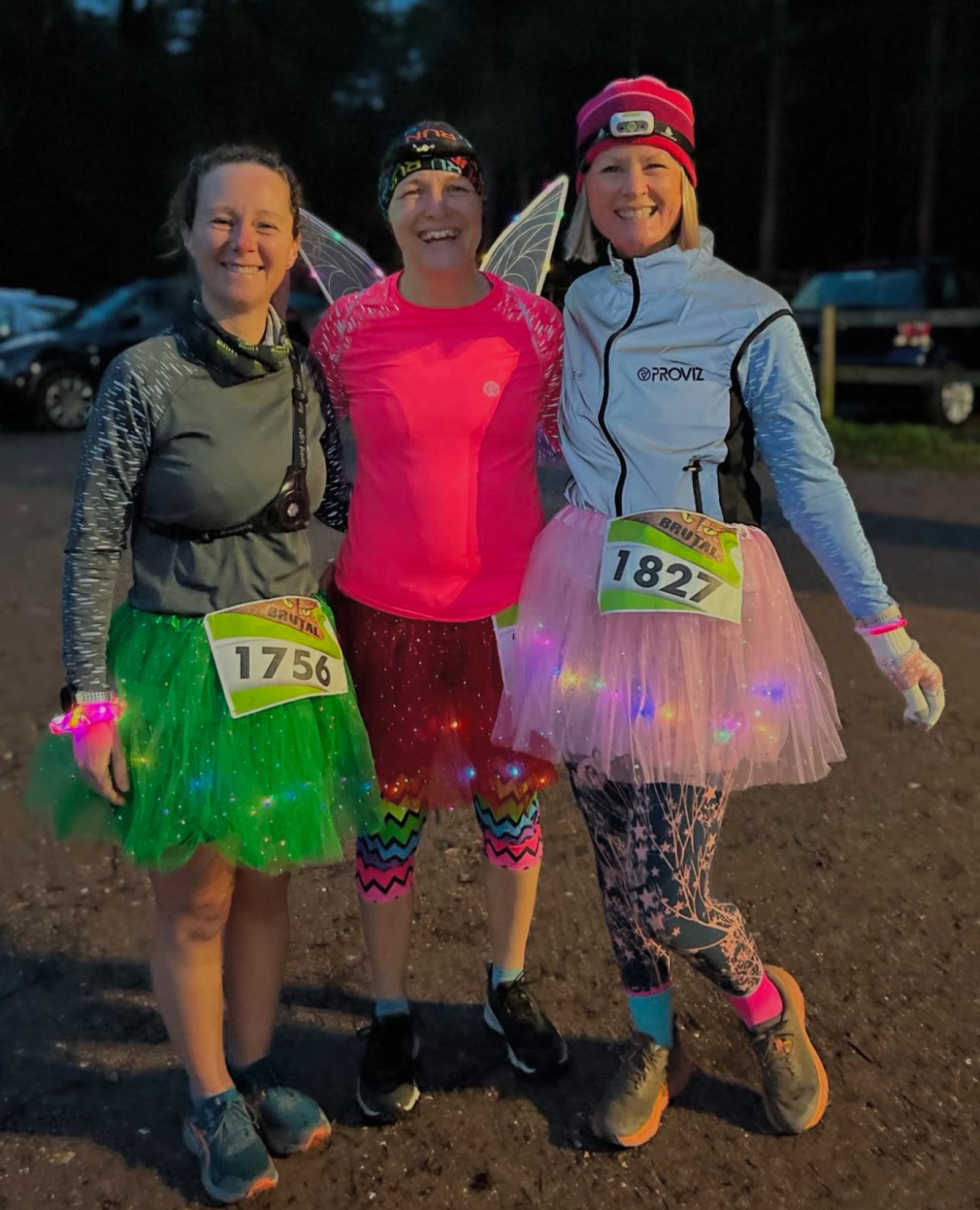 Three people in colorful running attire and tutus pose together, smiling. They wear numbered race bibs and glowing accessories. The background suggests an outdoor event, possibly early morning or evening.