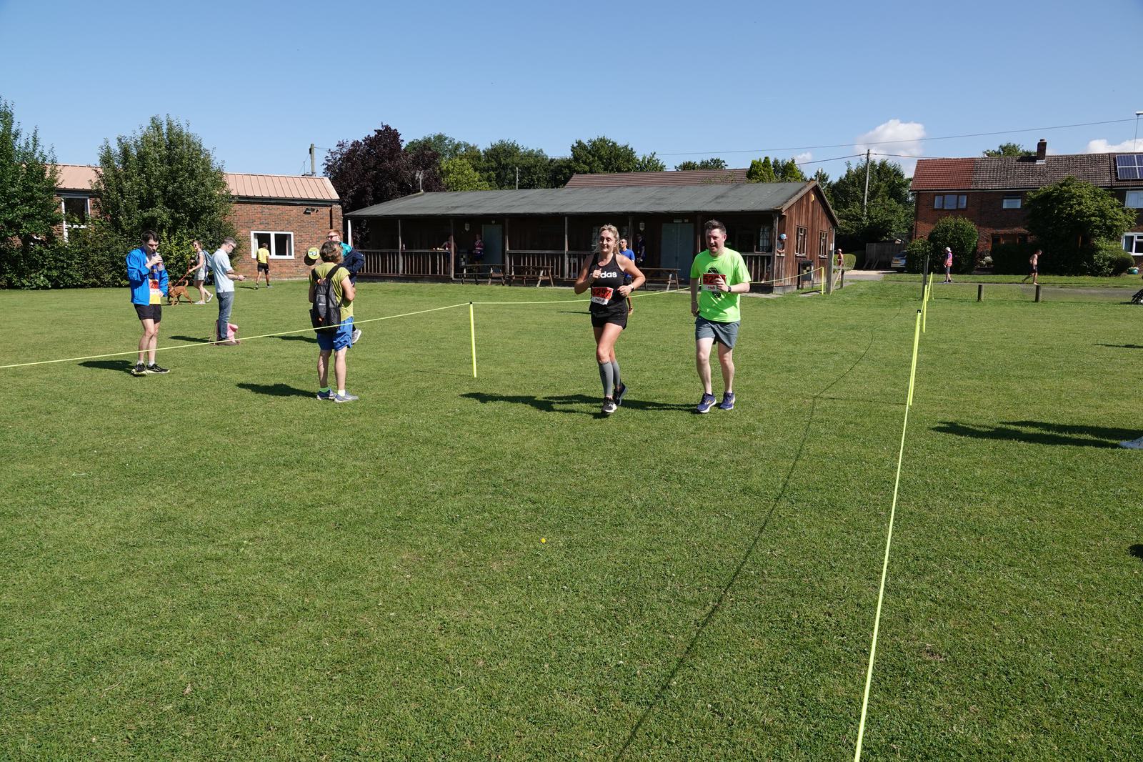 A group of runners approaches the finish line on a grassy field. A man in a green shirt and a woman in a blue shirt run together, hand in hand. Spectators and volunteers are nearby, with buildings and trees in the background.