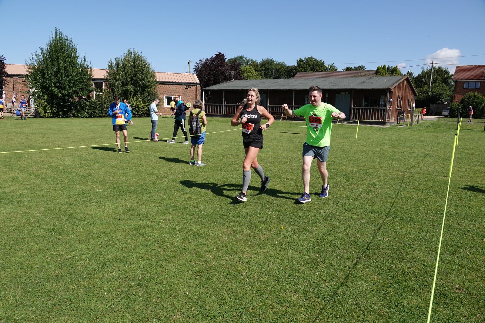 Two runners crossing a grassy finish line at an outdoor event. The man on the right raises his arms in celebration, and the woman next to him smiles. In the background, a few people stand near a building and trees under a clear blue sky.