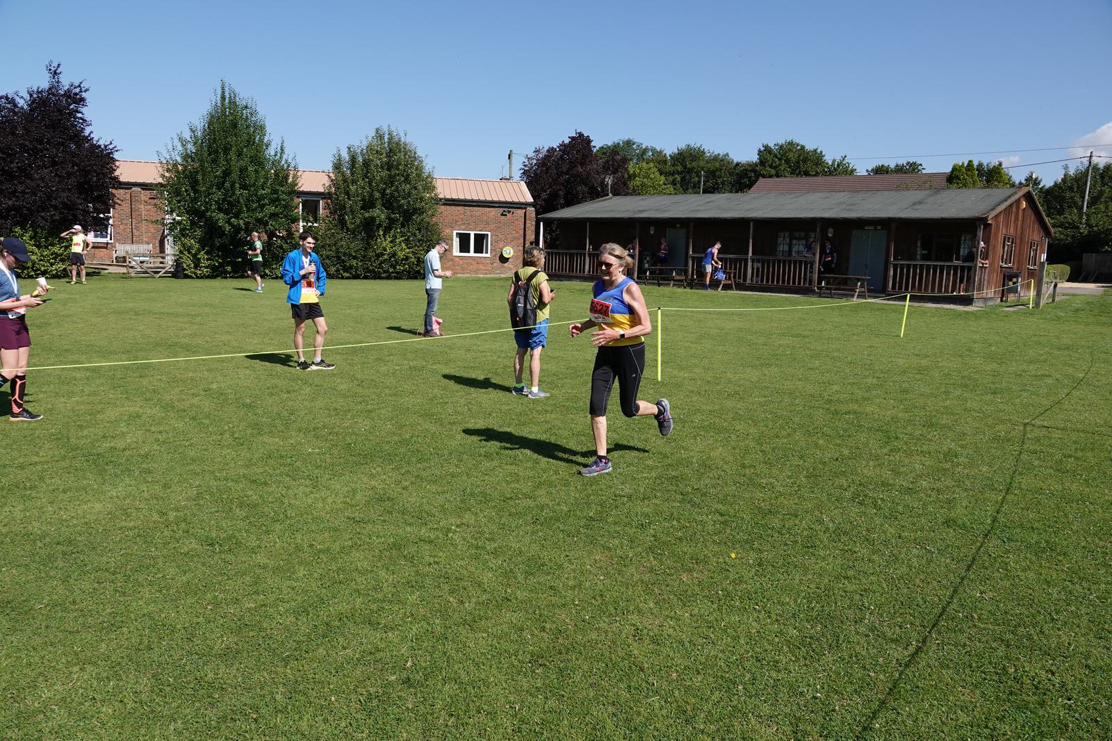 A woman in athletic wear runs across a grassy field during a race, while spectators and other participants are visible in the background. A small building and trees line the perimeter under a clear blue sky.
