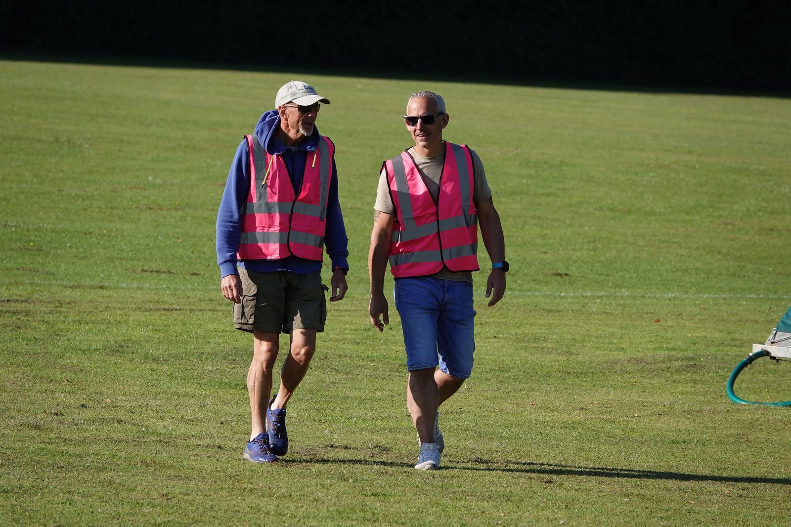 Two people wearing pink reflective vests are walking on a grassy field. One is wearing a cap and sunglasses, while the other sports shorts and sunglasses. They appear to be engaged in conversation on a sunny day.