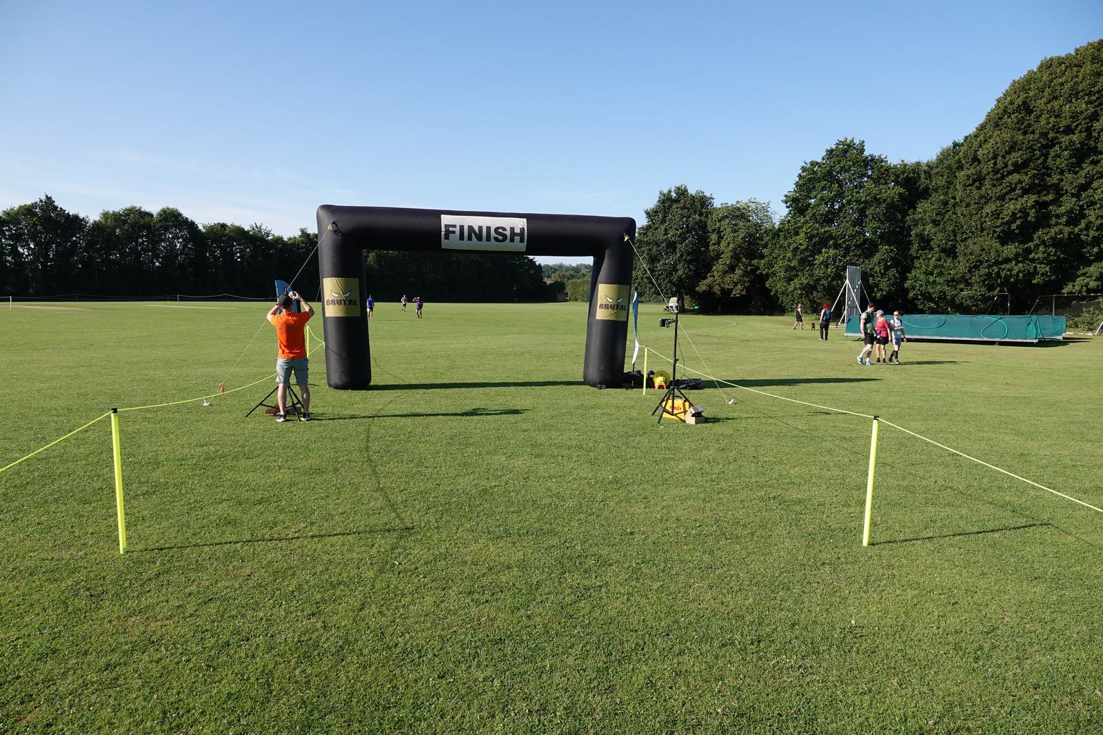 A grassy field with a black inflatable finish line arch labeled "FINISH." A person in an orange shirt stands to one side adjusting equipment. People are seen in the background near trees and a folded green structure.