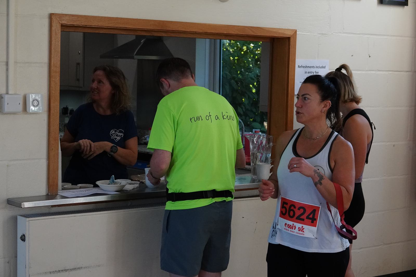 A runner with a red race number 5624 and a white tank top pauses near a refreshment window. A woman stands behind the counter, while a man in a bright green shirt faces away, wearing a belt pack.