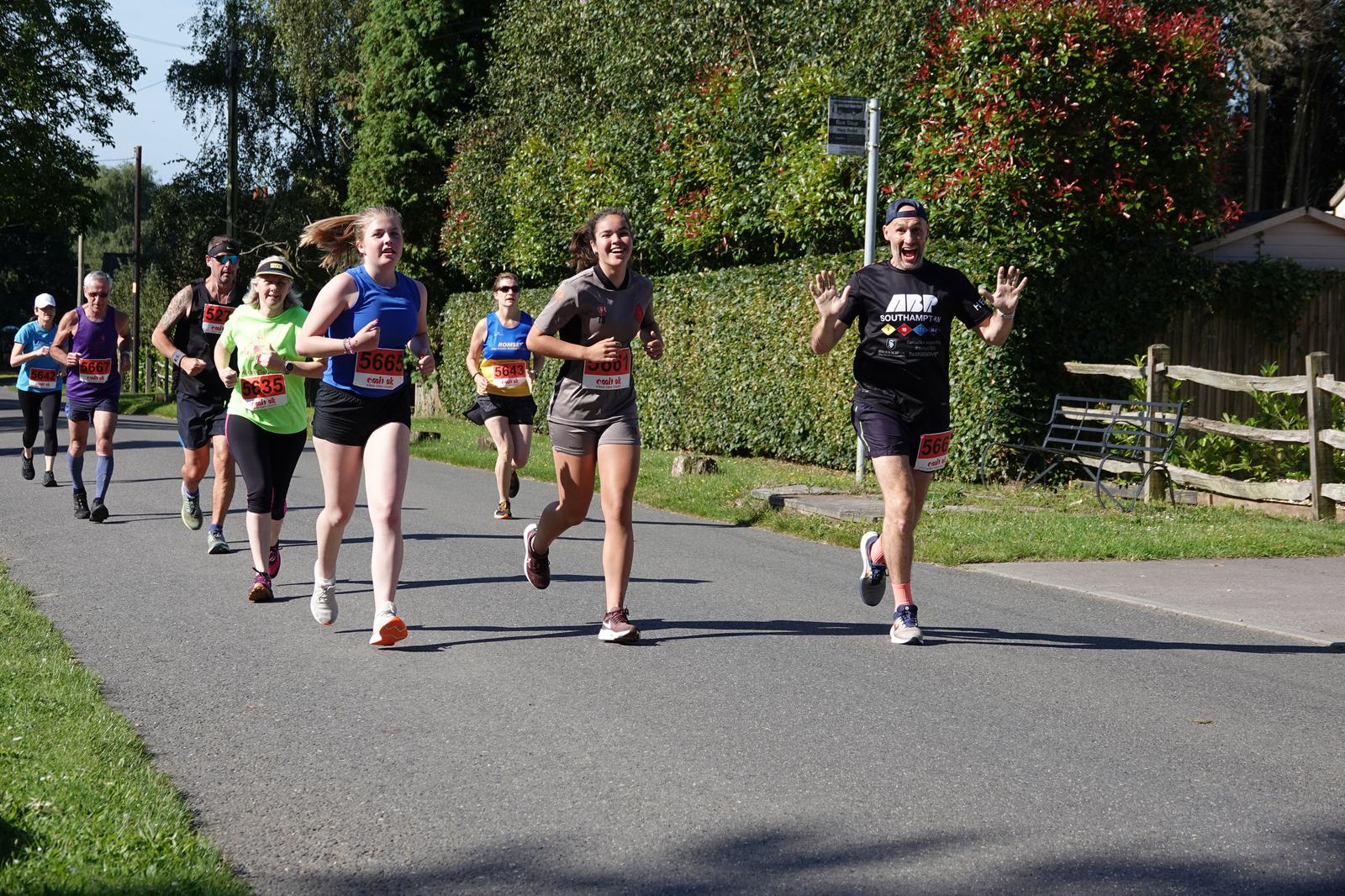 A group of runners participating in a marathon on a sunny day, with trees and a wooden fence in the background. Some runners smile and wave at the camera while wearing race bibs and casual sport attire.