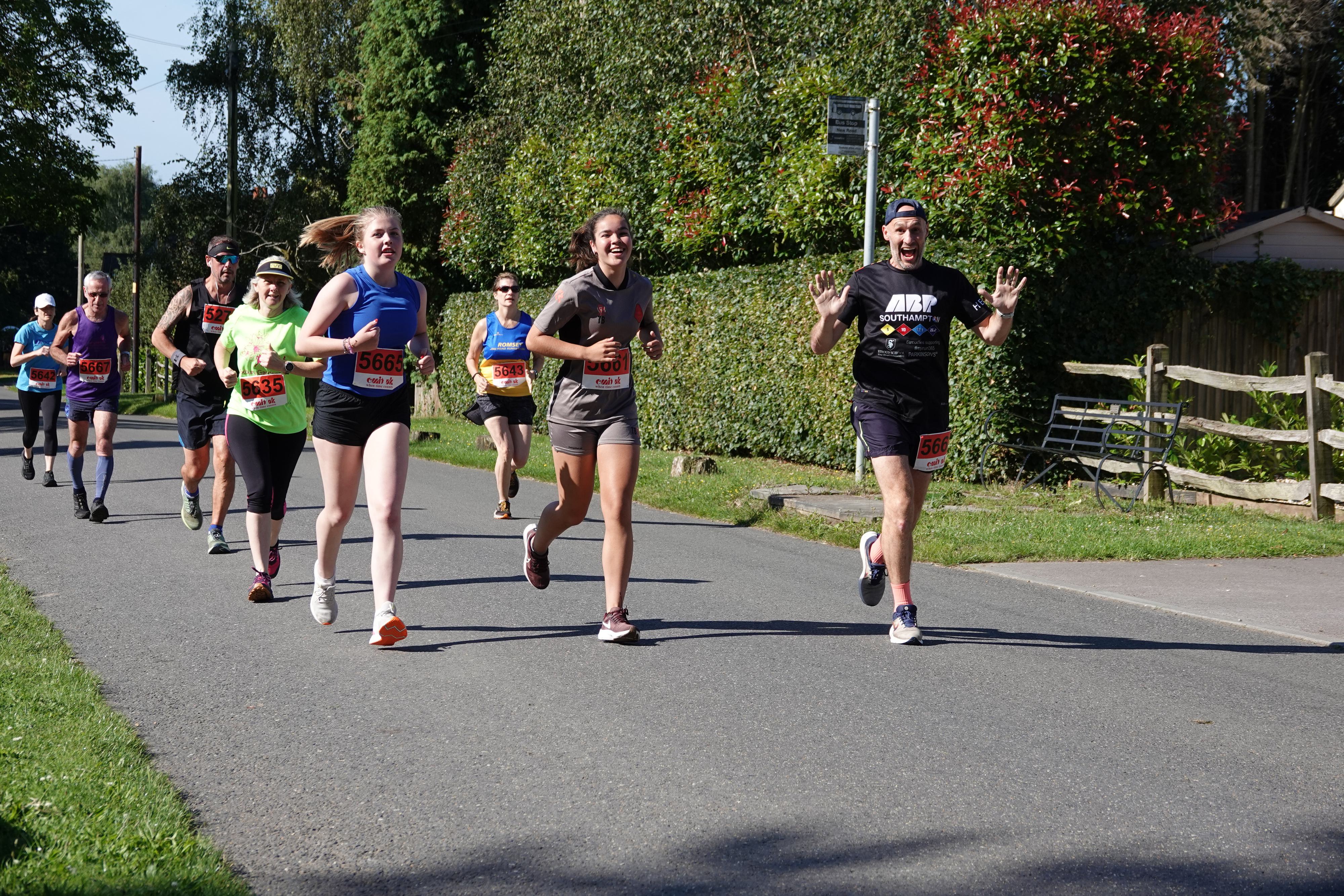 A group of runners participating in a marathon on a sunny day, with trees and a wooden fence in the background. Some runners smile and wave at the camera while wearing race bibs and casual sport attire.