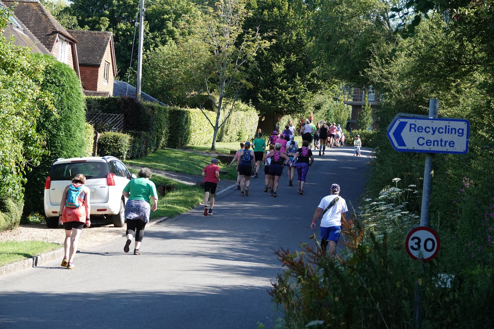 A group of people participating in a running event along a suburban road lined with trees and houses. Some are walking or jogging. A sign for a recycling center and a 30 mph speed limit sign are visible.