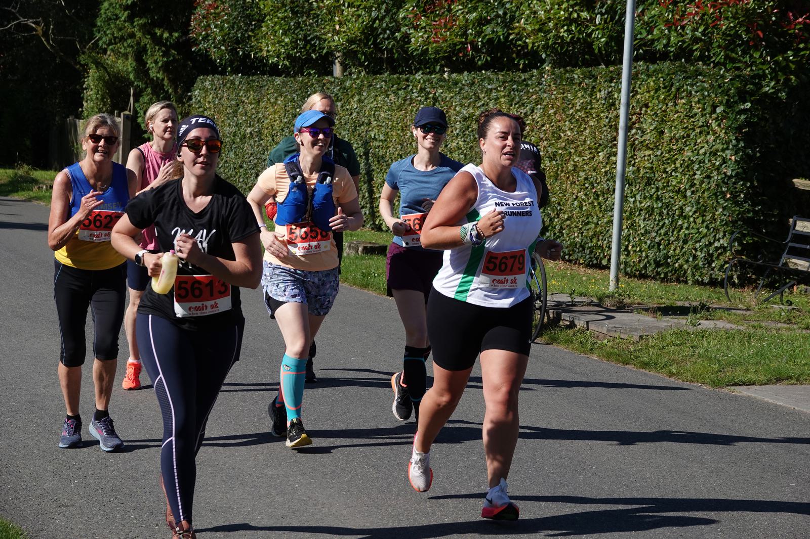 A group of runners in colorful athletic wear participate in an outdoor race. They are jogging on a paved road with green bushes lining the path under a sunny sky. Some runners wear sunglasses and hats.