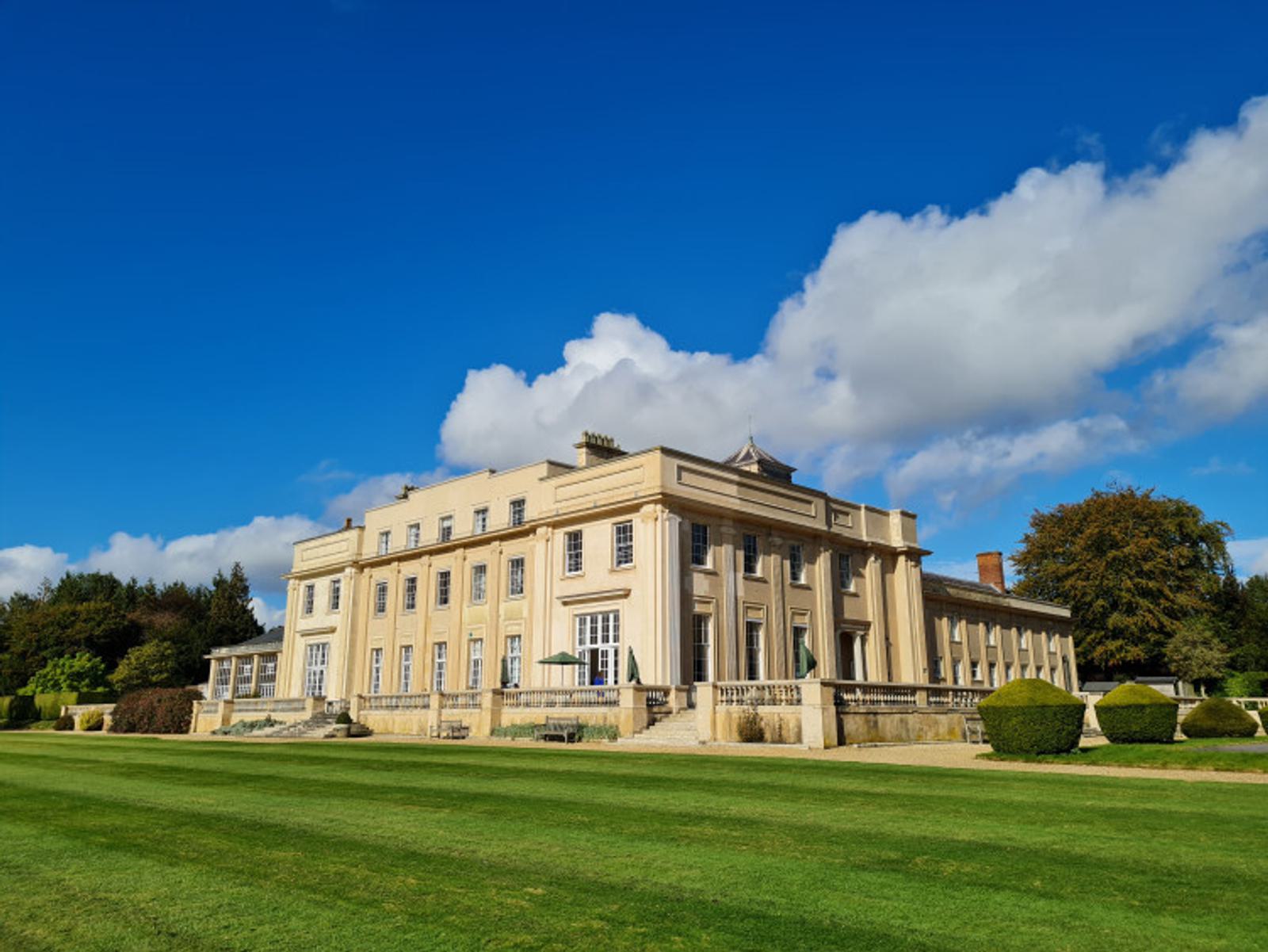 A large, historic mansion with cream-colored walls and numerous windows stands under a bright blue sky with scattered clouds. The manicured lawn and trimmed hedges surround the building, enhancing its grandeur.