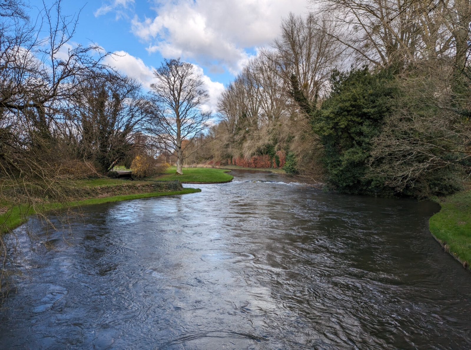 A gently flowing river curves through a park with bare trees and green grass under a partly cloudy blue sky. Bushes and leafless branches line the riverbanks.