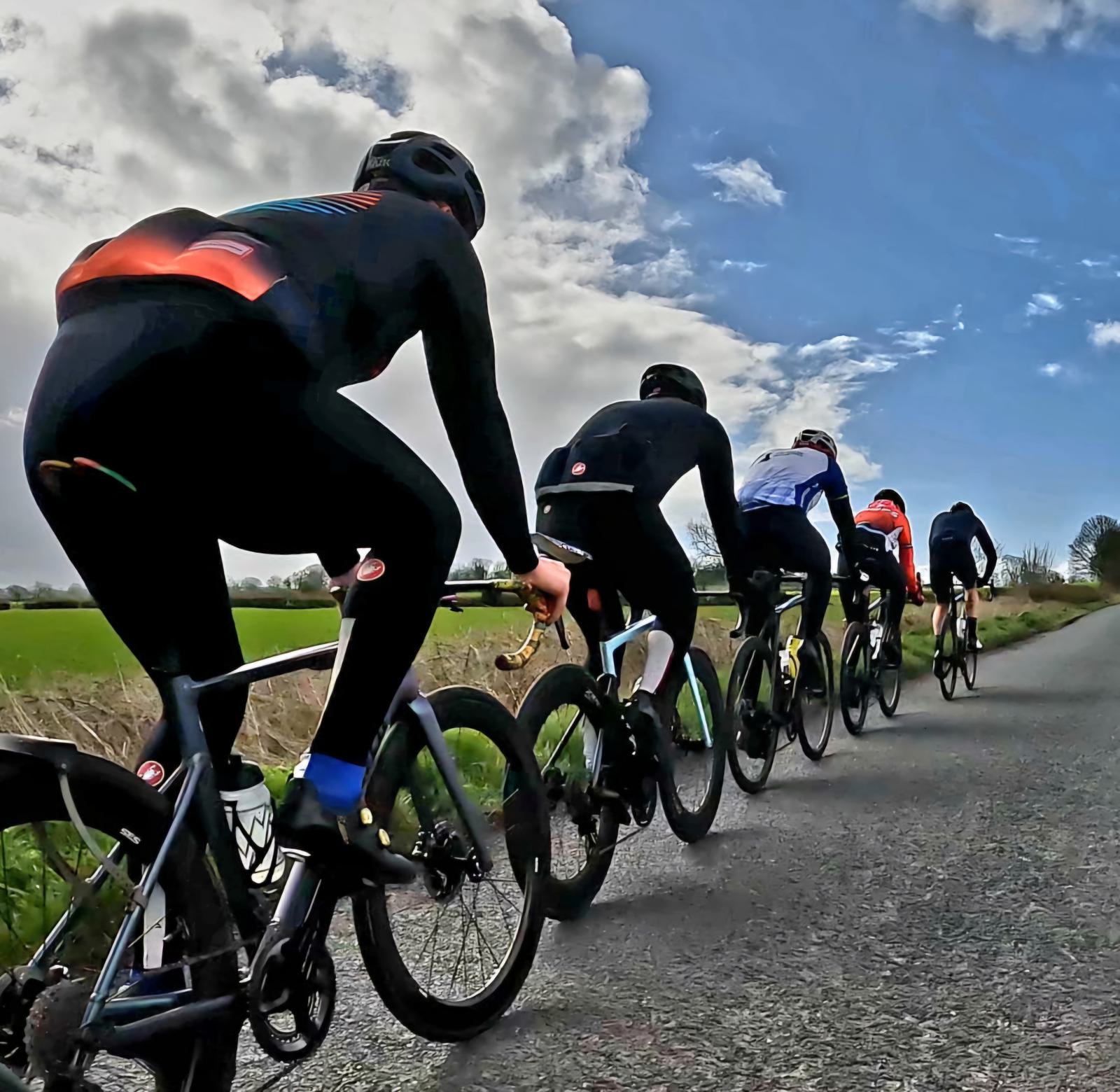 A group of six cyclists dressed in athletic gear are riding their bikes on a rural road. They are aligned in a single file under a partly cloudy sky, with green fields on either side of the road.