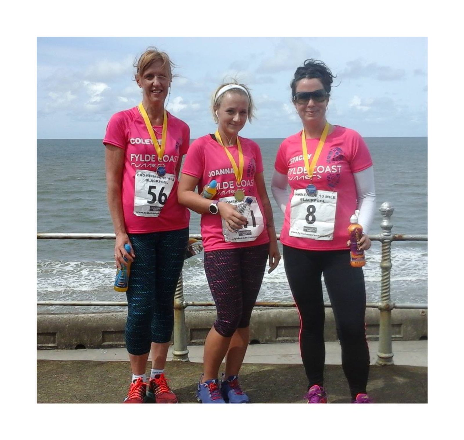 Three women in pink shirts and leggings stand on a seaside promenade after a race, wearing medals and holding drinks. They are smiling and the ocean is visible in the background under a partly cloudy sky.