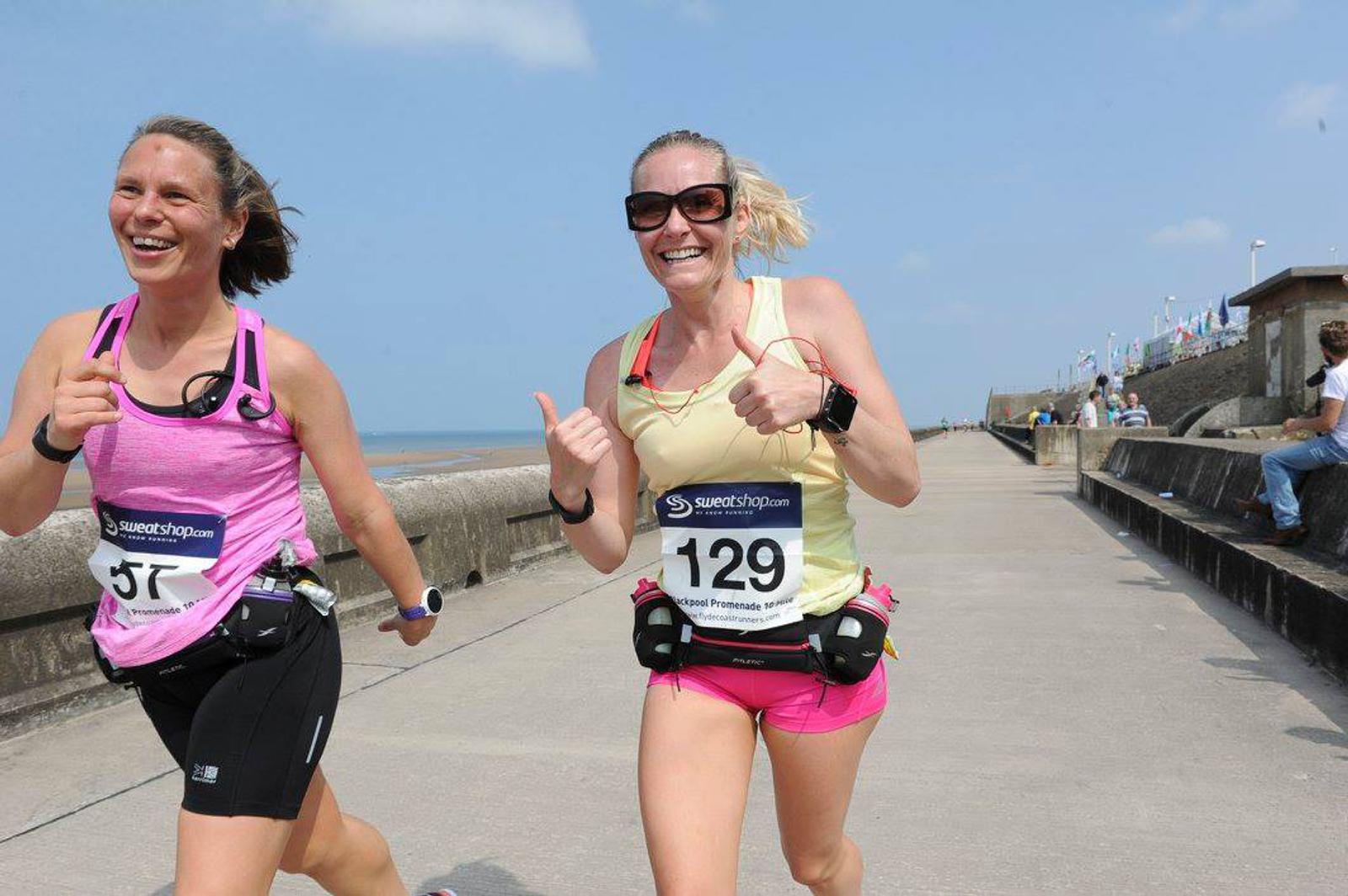 Two women are running on a seaside pathway. Both are wearing athletic gear with race numbers, smiling as they run. The sky is clear and the sea is visible in the background, with people walking along the promenade.