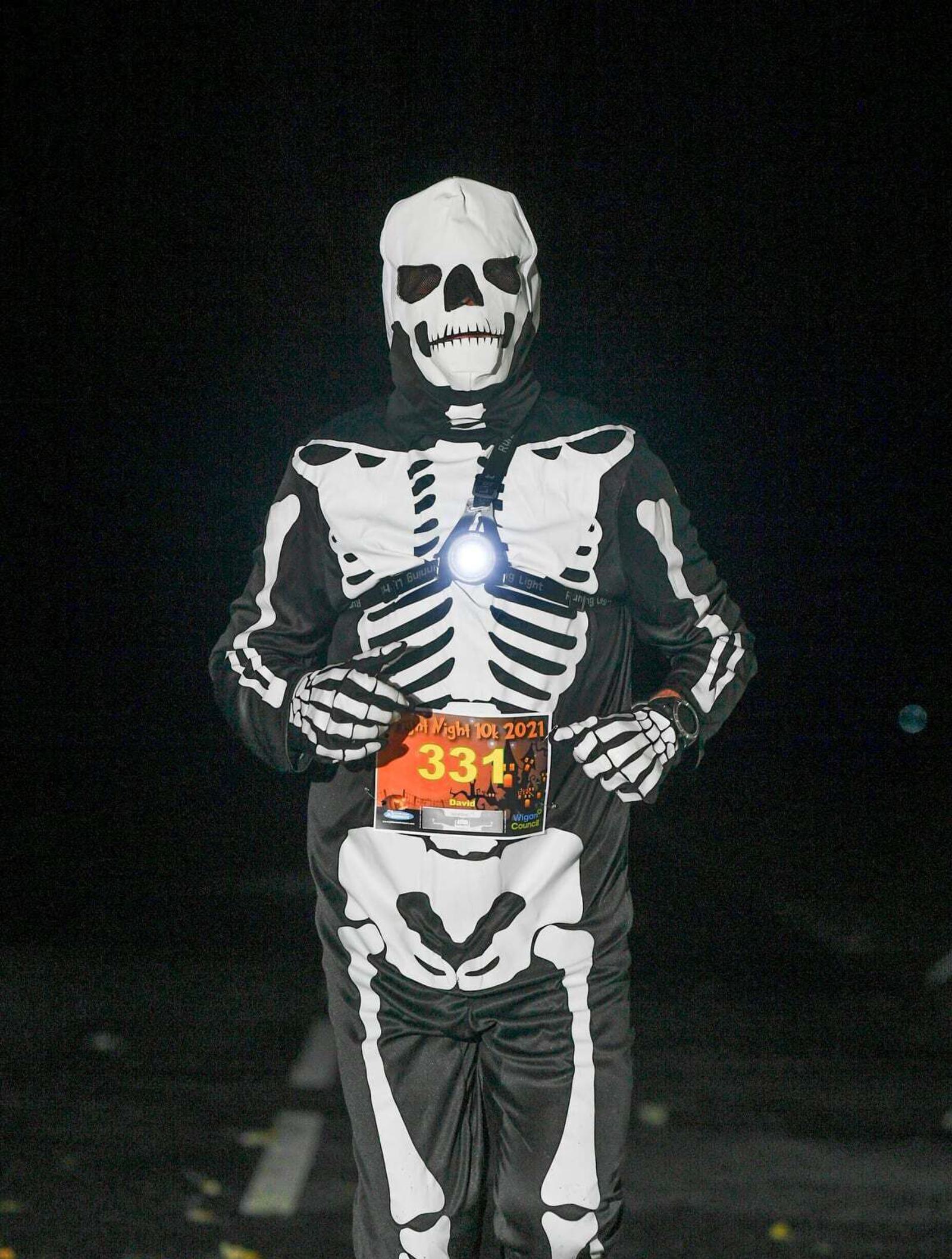 A person dressed in a skeleton costume is running at night. They wear a headlamp and have a race bib with number 331 on their chest. The background is dark, emphasizing the illuminated skeleton design.