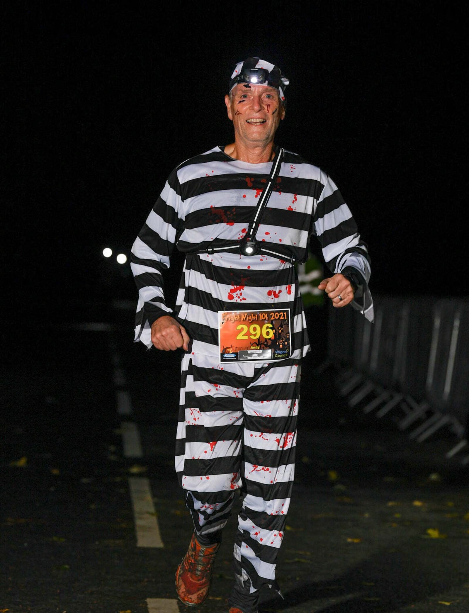 A person dressed in a striped prisoner costume, decorated with fake blood, runs on a dark road. They're wearing a race number "296." Logos on the image include Fylde Coast Runners, Haigh Woodland Park, and Wigan Council.