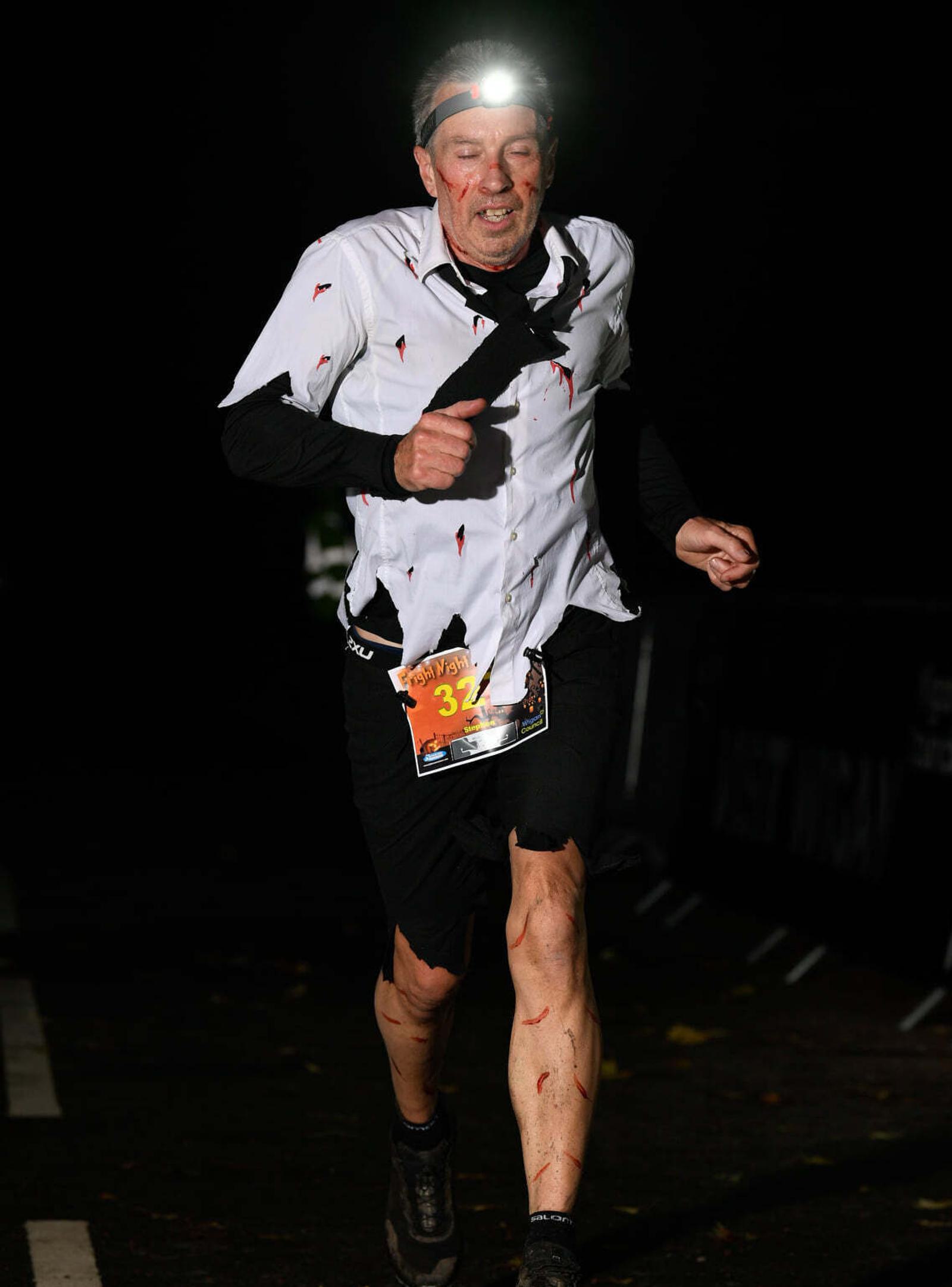 A man wearing a torn costume and a headlamp runs in a night race. The background is dark, suggesting an evening event. Logos at the bottom indicate event sponsors: Fylde Coast Runners, Haigh Woodland Park, Wigan Council, etc.