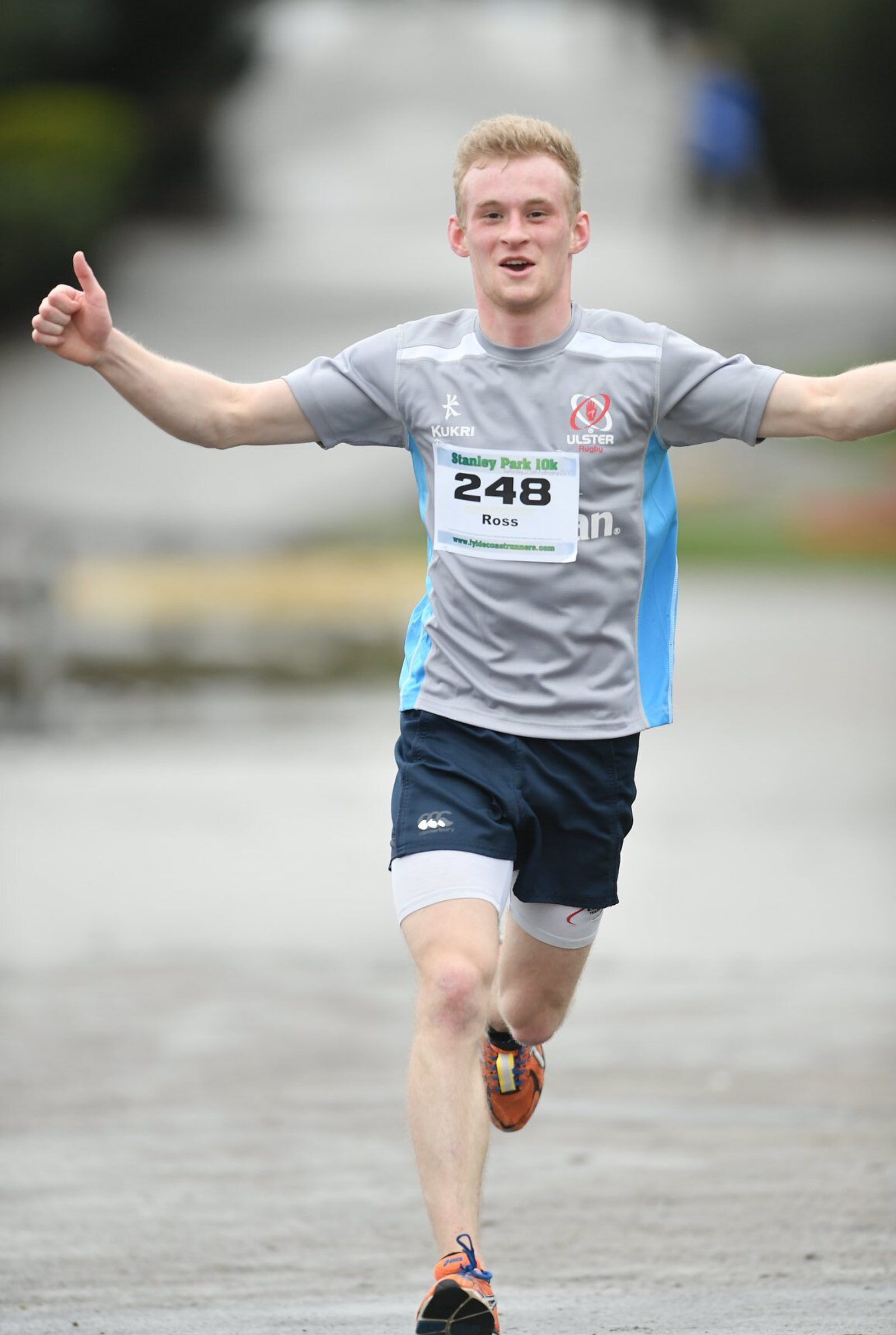 A runner in a grey and blue shirt crosses the finish line with arms raised, wearing a race bib labeled "248" with the name "Ross" visible. The background is a blurred park setting.