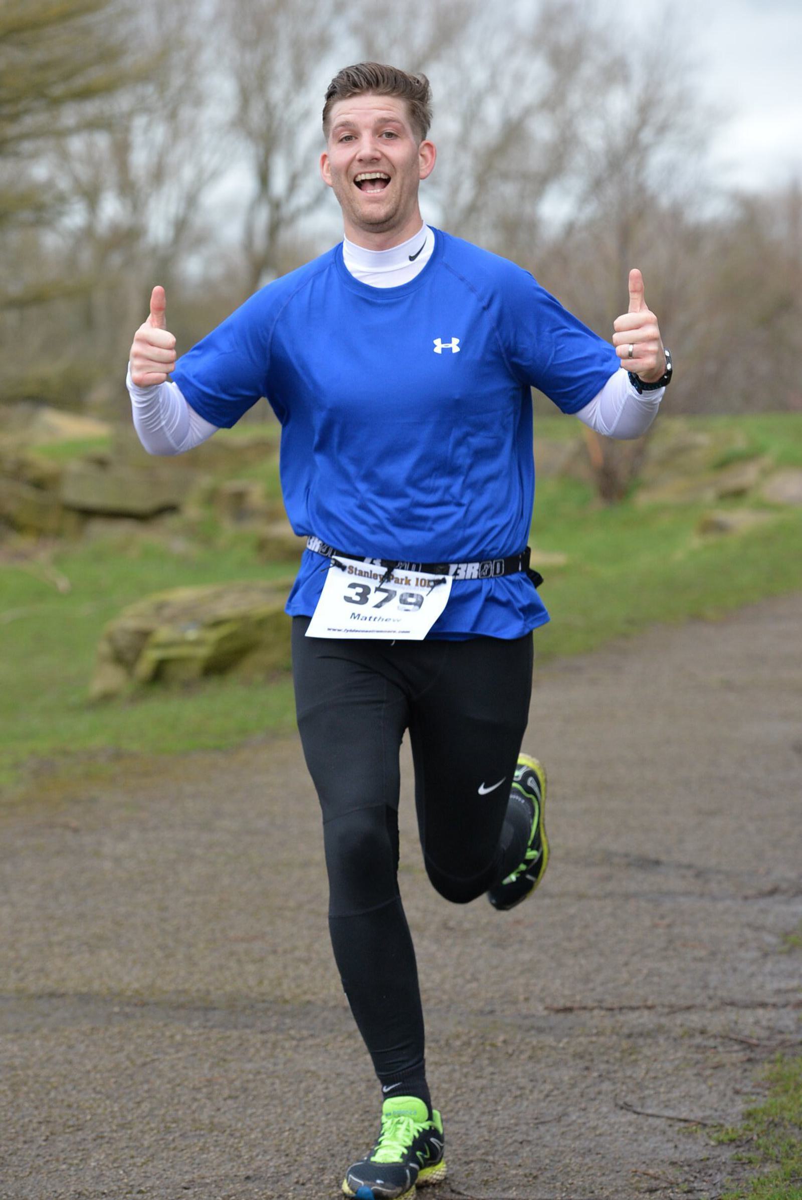 A person in a blue shirt and black leggings is running outdoors on a path, giving a thumbs-up with both hands. They are smiling, and a race bib with the number 329 is visible. Trees and grass are in the background.